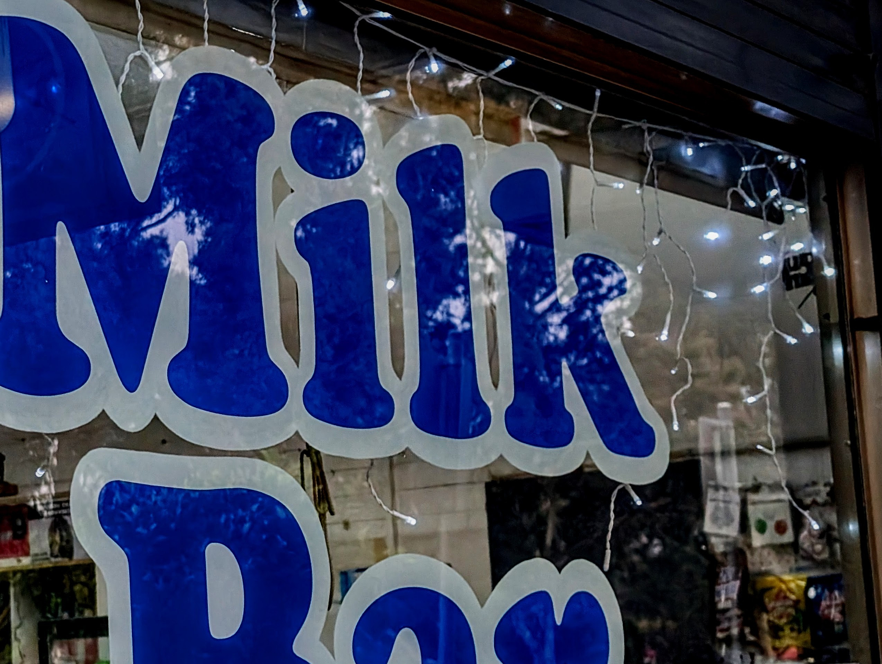Large blue lettering spelling Milk Bar is displayed on a window adorned with string lights.