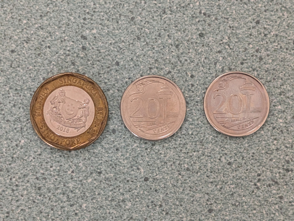 Three Singapore coins are placed in a row on a textured surface, with one 50 cent coin from 2012 and two 20 cent coins.