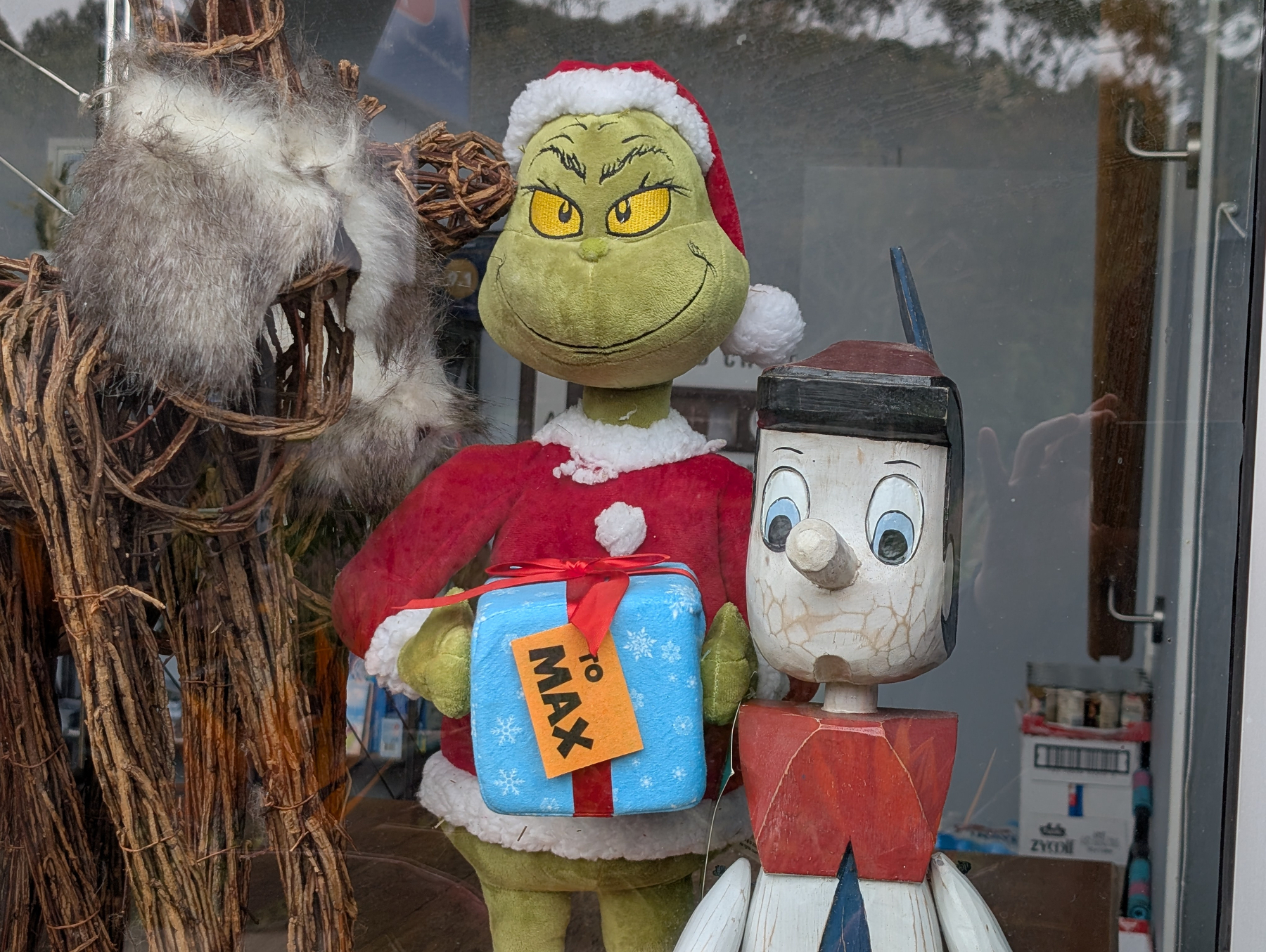 A Grinch figure holding a gift labeled To Max stands next to a wooden puppet-like figure in a window display.
