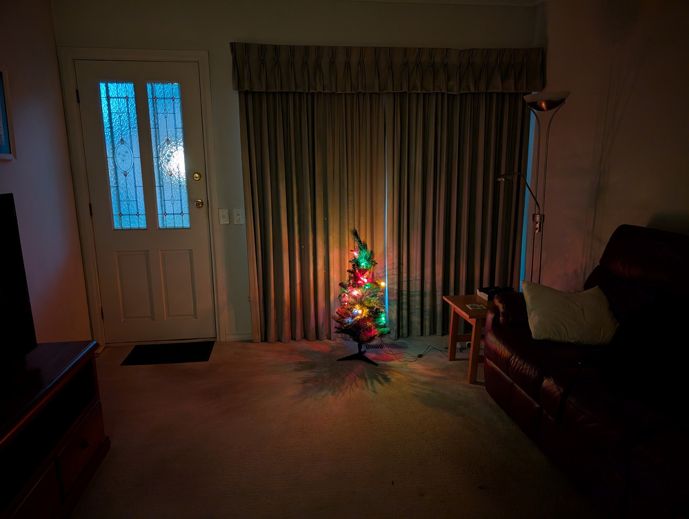 A small, brightly lit Christmas tree is set up in a dimly lit living room next to a couch and in front of closed curtains.