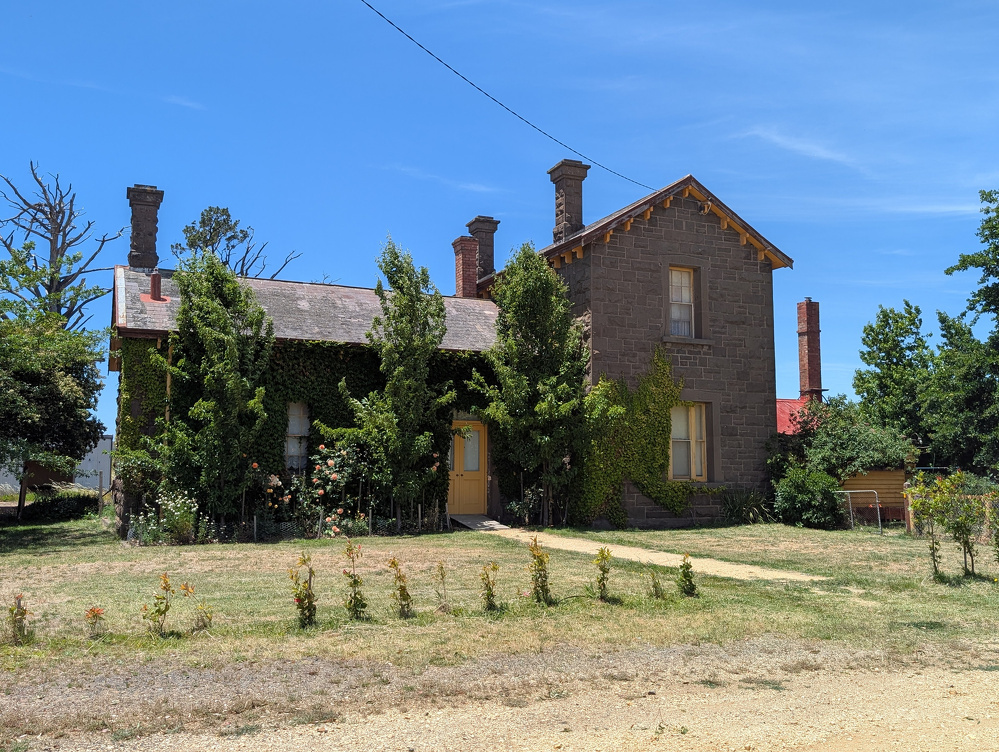 A rustic, ivy-covered stone house with chimneys stands surrounded by trees on a sunny day.