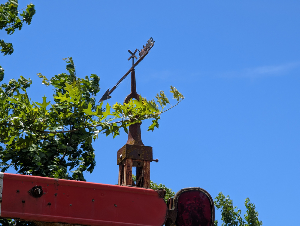 A weather vane with a rusted arrow and directional cross sits atop a red structure, surrounded by green leaves and set against a clear blue sky.