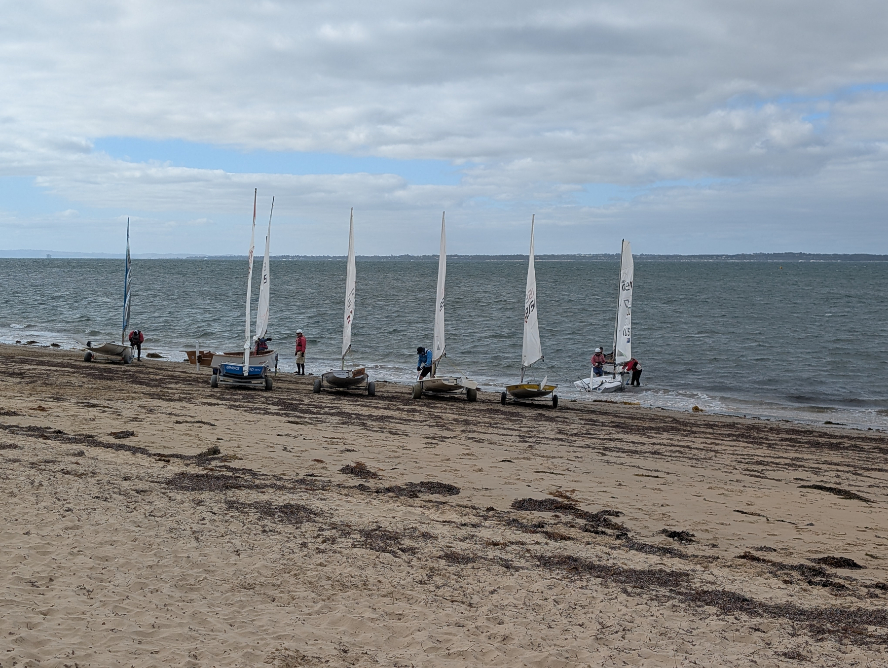 A group of people are preparing small sailboats along a sandy beach under a cloudy sky.