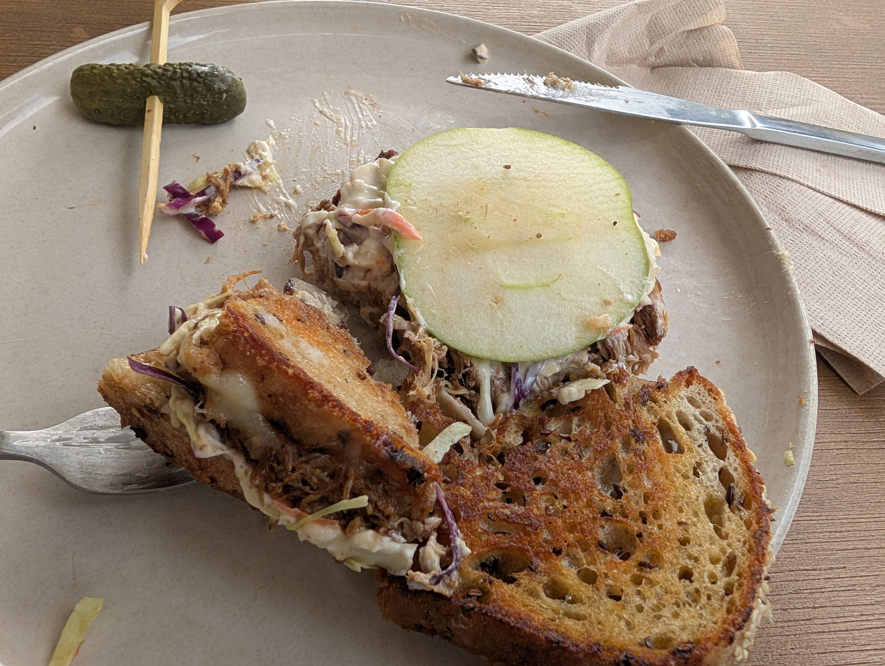 A partially eaten sandwich with melted cheese, meat, coleslaw, and a slice of green pear sits on a plate alongside a small pickle on a toothpick.