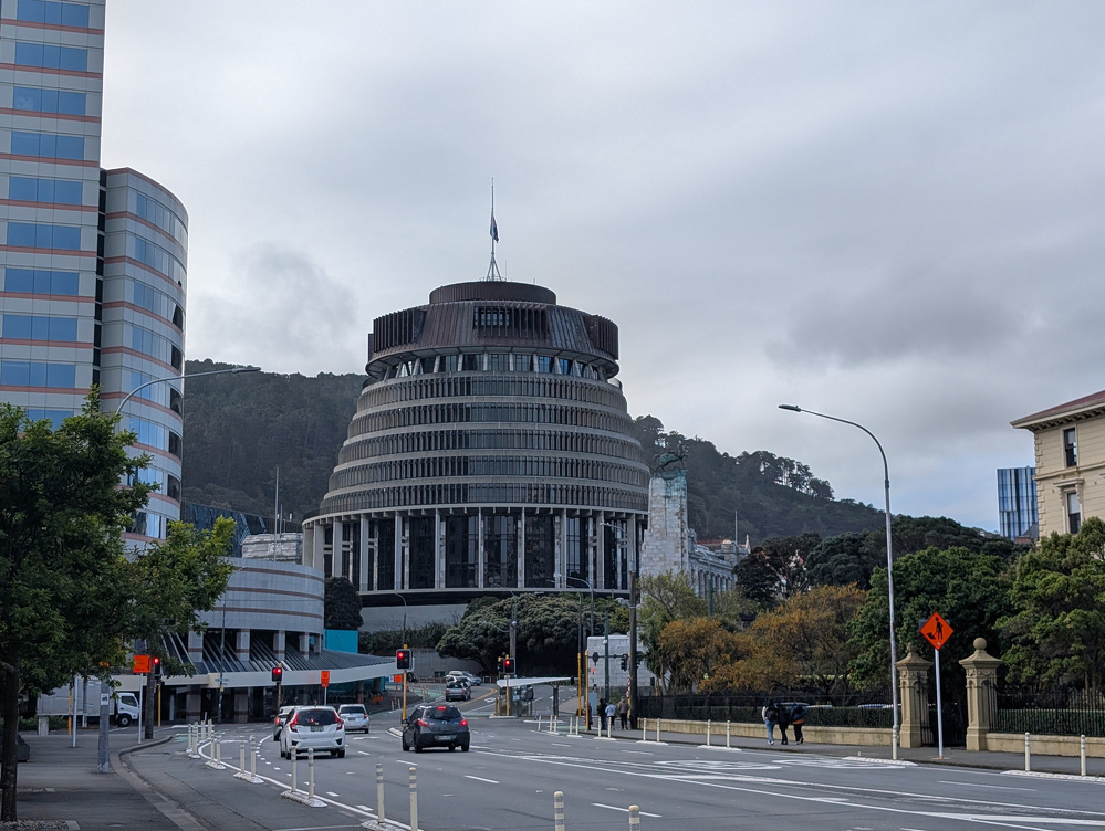 The New Zealand Parliament, known as the Beehive. A distinctive round building with horizontal lines and a dark top, located at the end of a street lined with trees and modern buildings.
