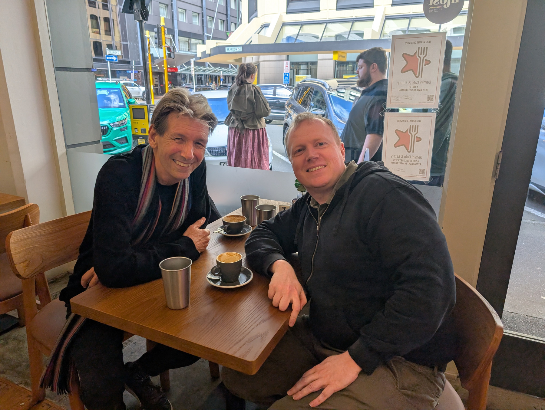 Auto-generated description: Two men are sitting at a wooden table inside a café, enjoying coffee and smiling at the camera.
