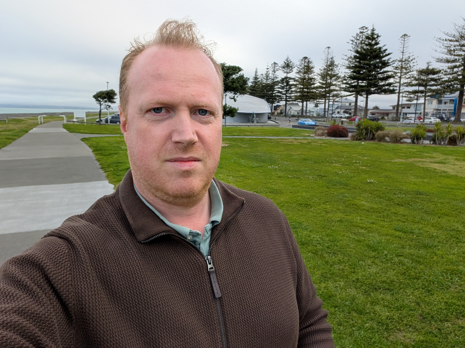 A man wearing a brown jumper stands on a pathway with a grassy area and trees in the background.