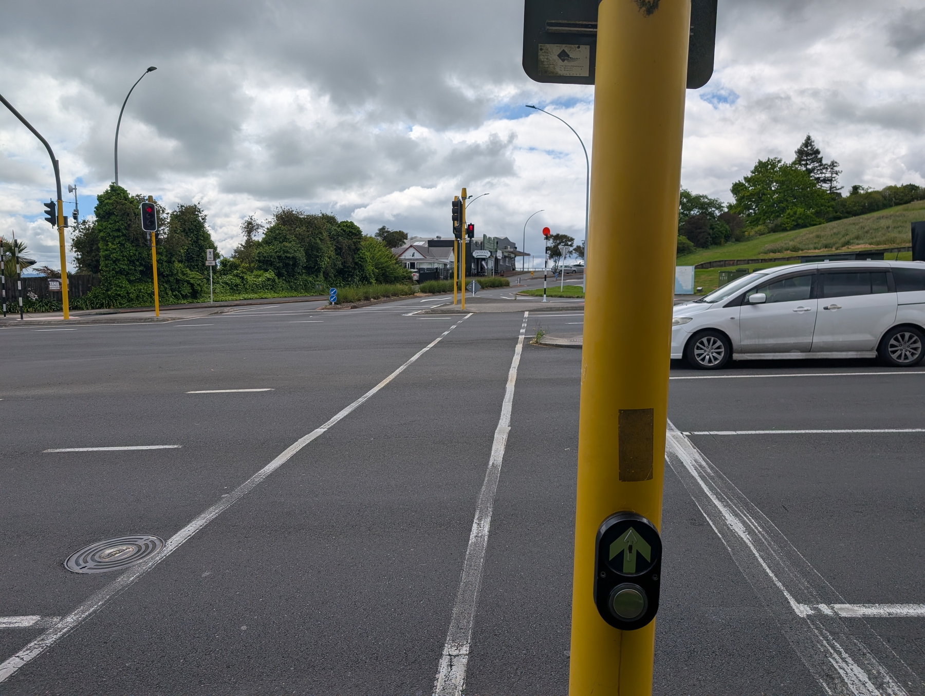 Auto-generated description: A pedestrian crosswalk button is mounted on a yellow pole at a traffic intersection with cars and traffic lights visible under a cloudy sky.