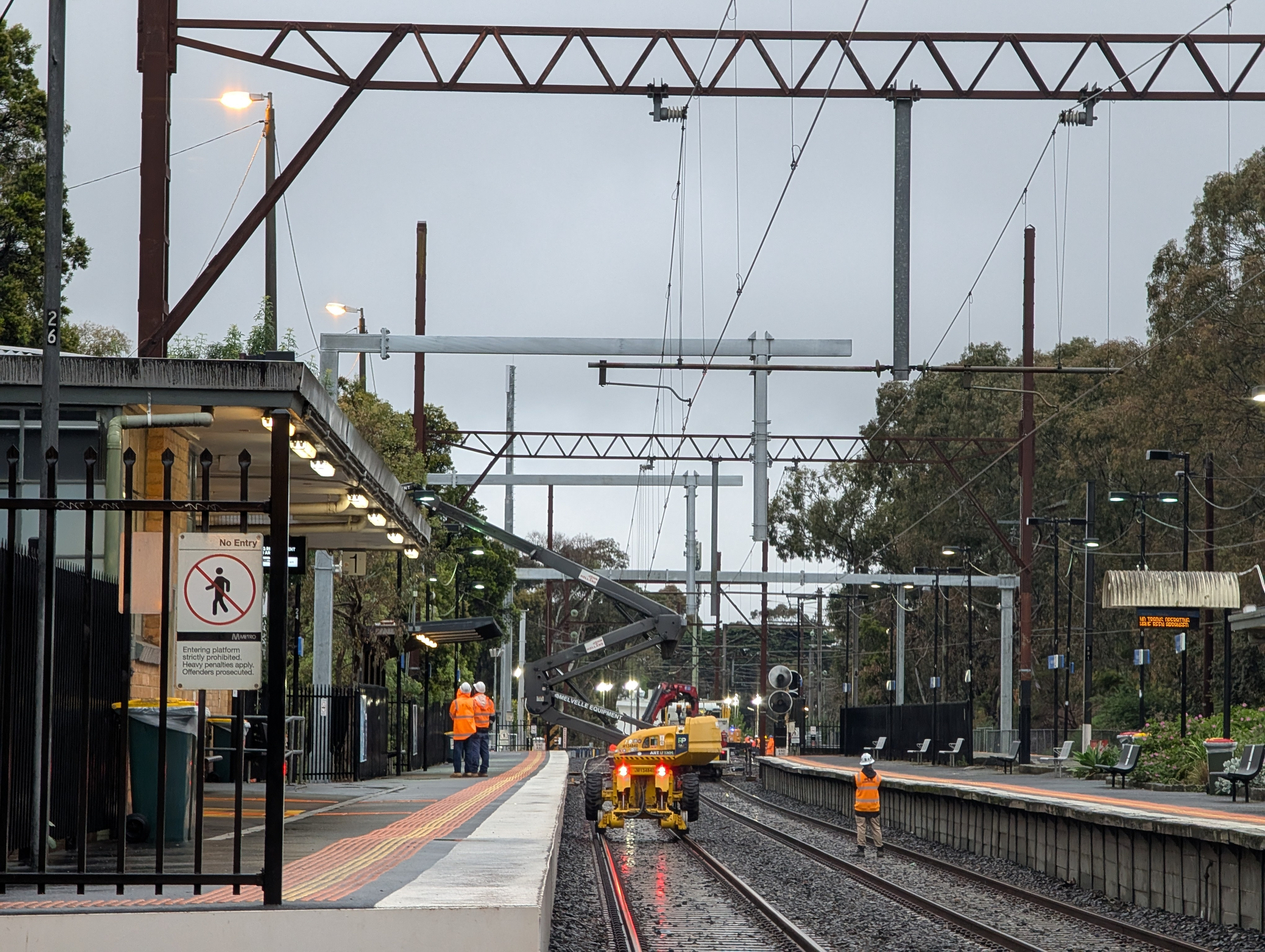 Workers in orange safety vests are conducting maintenance on a railway track at a train station during overcast weather.