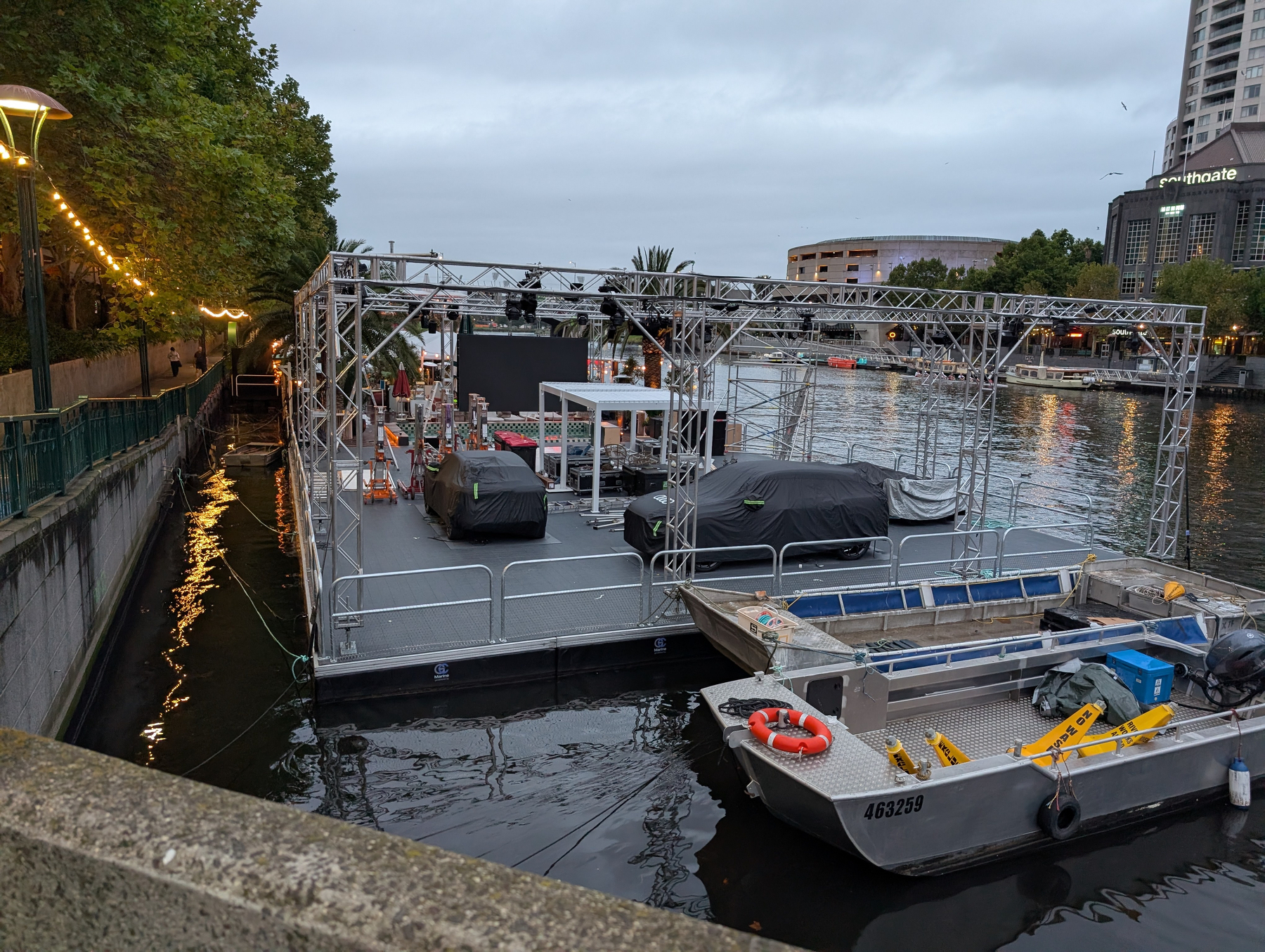 A floating platform with equipment and covered items is anchored next to a dock, with trees and buildings in the background.