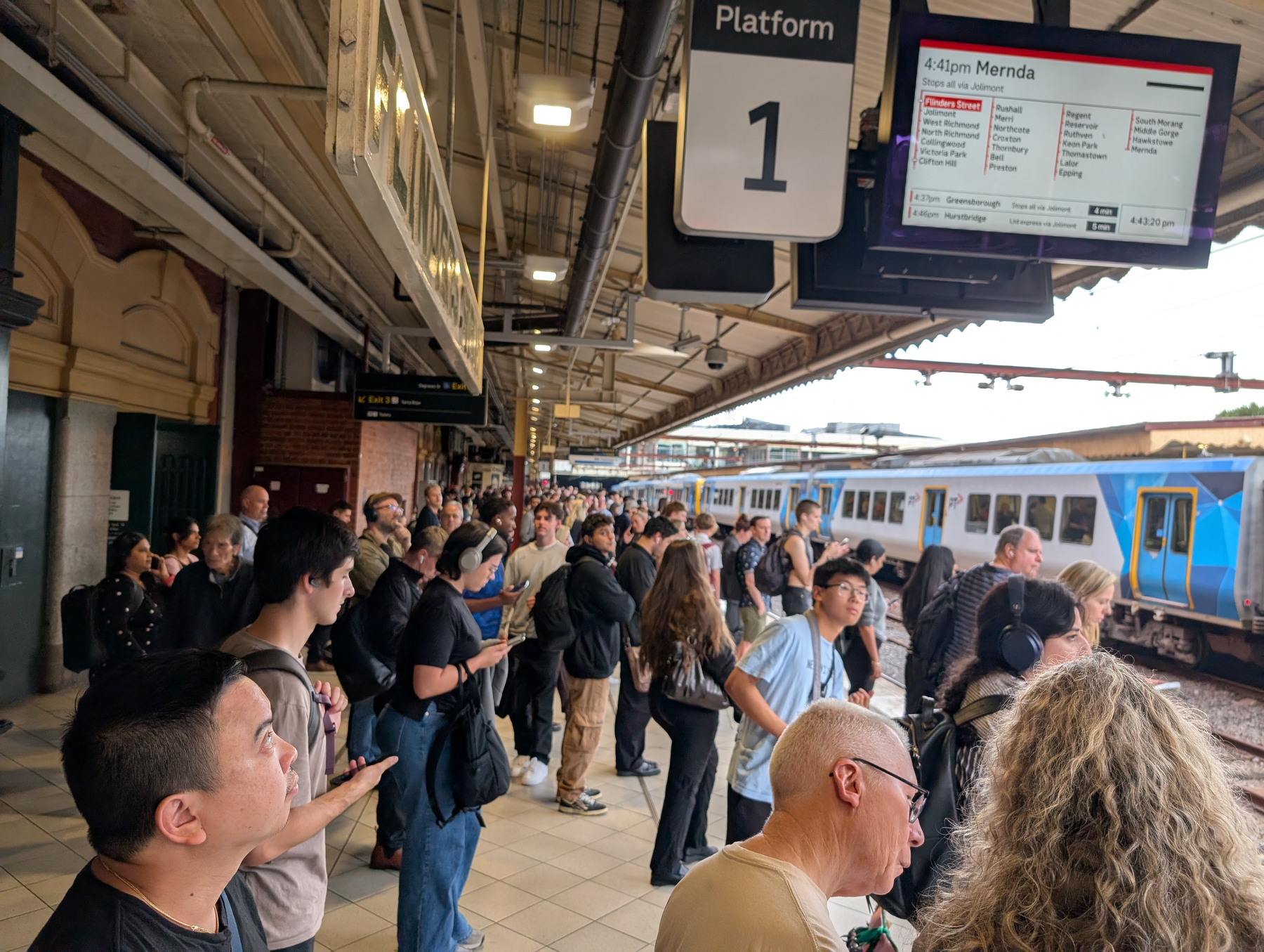 A crowd of people is waiting on a train platform labeled 1 with a digital sign displaying train information above.