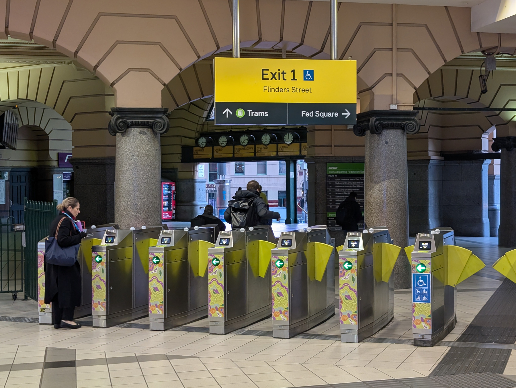 A group of people are passing through the turnstiles at a train station exit labeled Exit 1 Flinders Street with directions to trams and Fed Square.