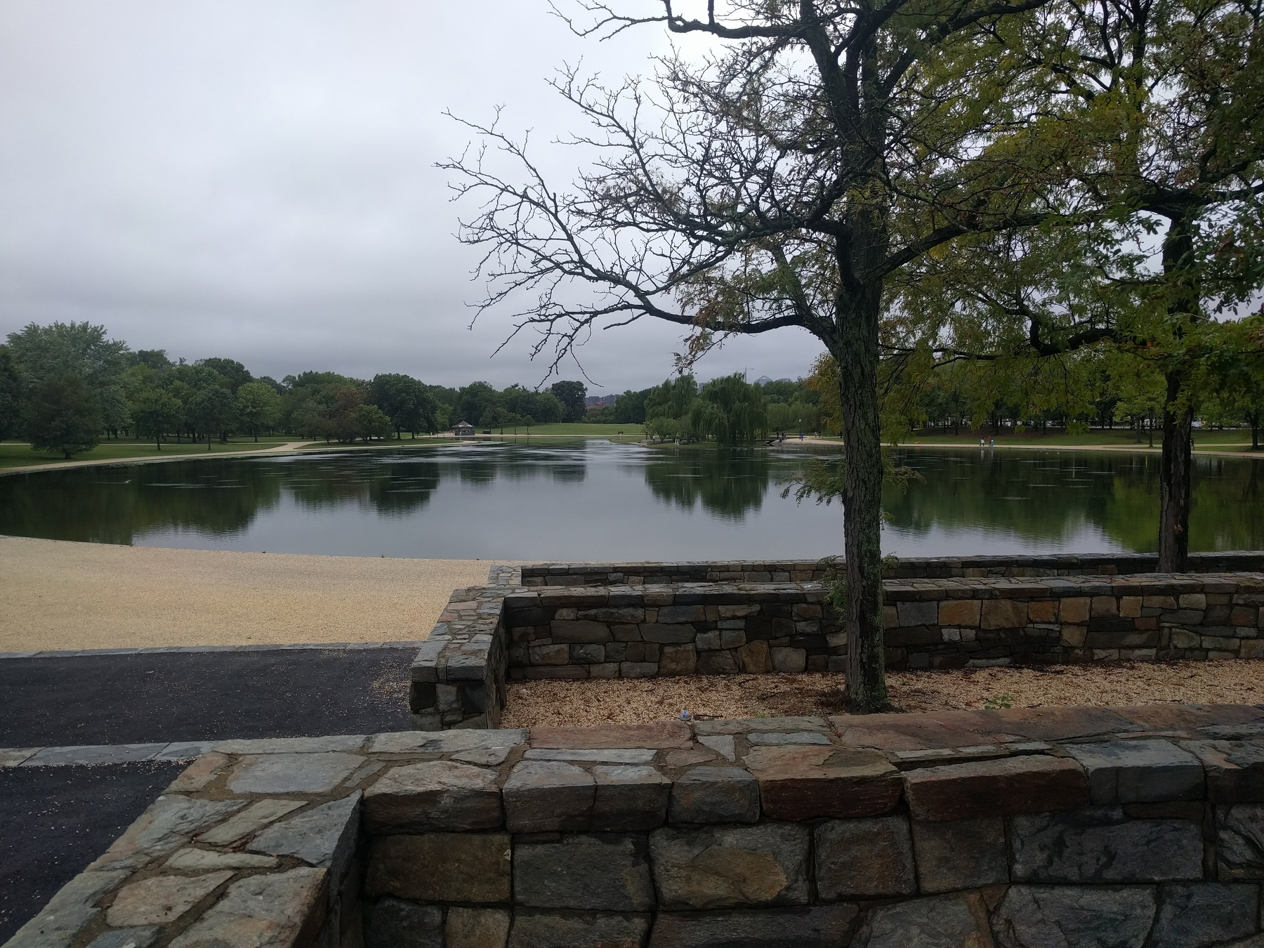 Auto-generated description: A calm lake is surrounded by trees and stone walls under a cloudy sky.