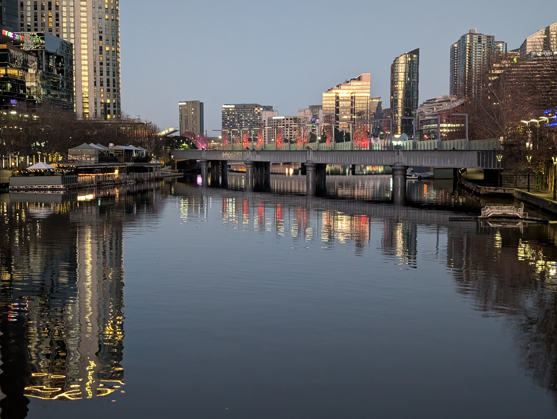 A serene urban river scene at twilight is framed by modern high-rise buildings and illuminated by colorful lights reflecting in the water.