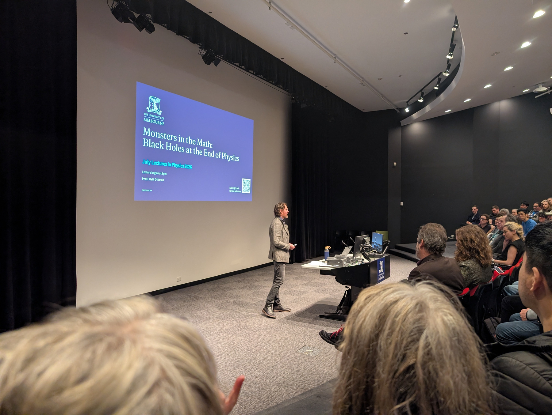 A speaker is giving a lecture titled Monsters in the Mantle: Black Holes at the End of Physics to an attentive audience in a lecture hall.