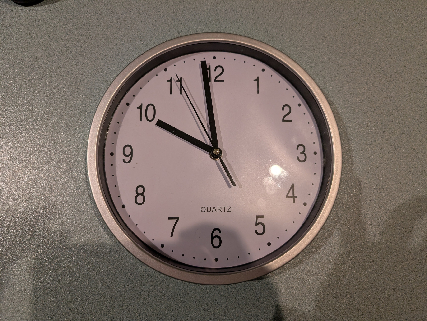 A round wall clock with an off-white face and a silver-coloured plastic rim shows the time as approximately 10:59.