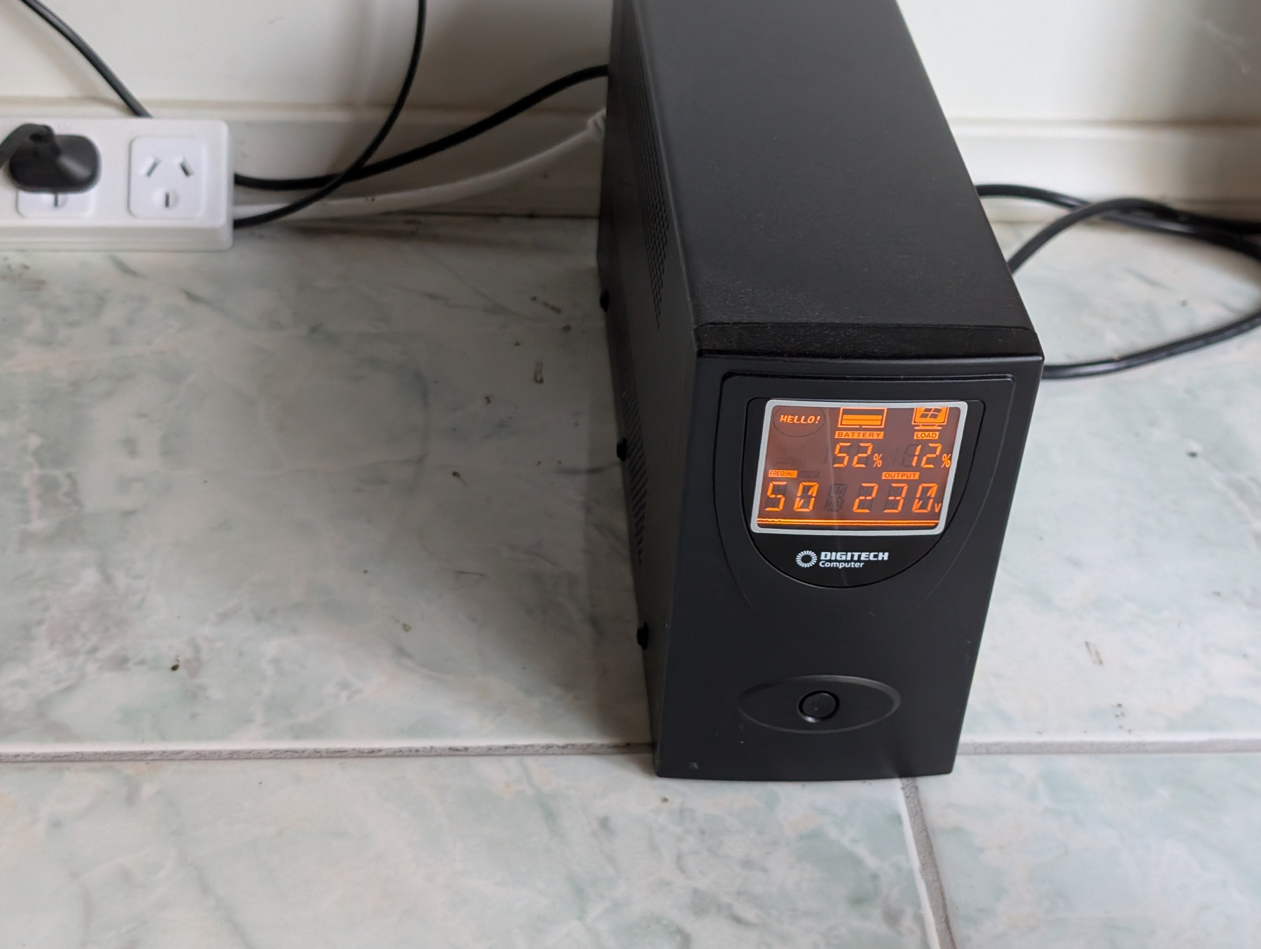 A black UPS (uninterruptible power supply) is positioned on a tiled floor, displaying various digital readings on its front panel.