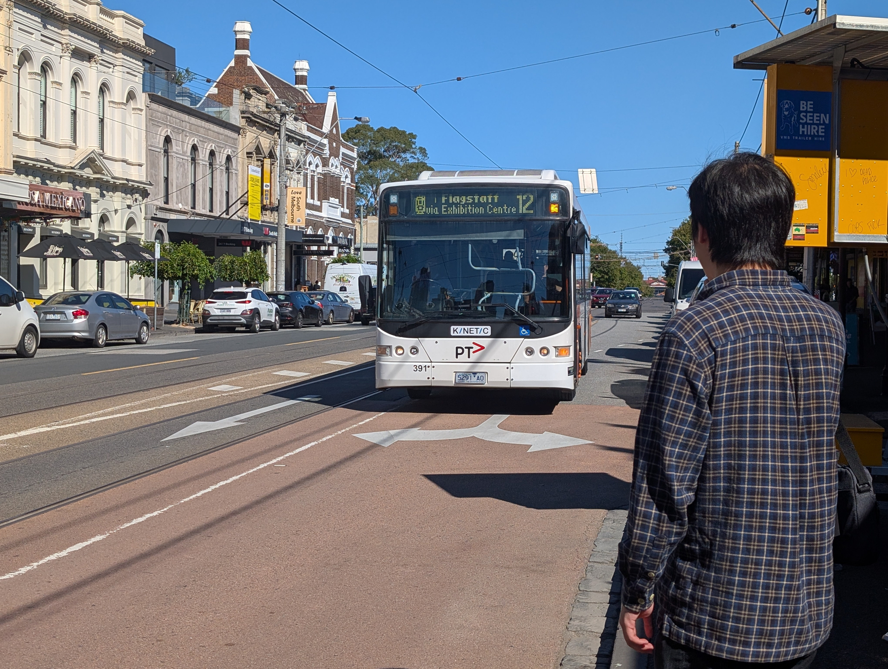 Auto-generated description: A person stands at a tram stop watching a bus approach on a busy street lined with historic buildings.