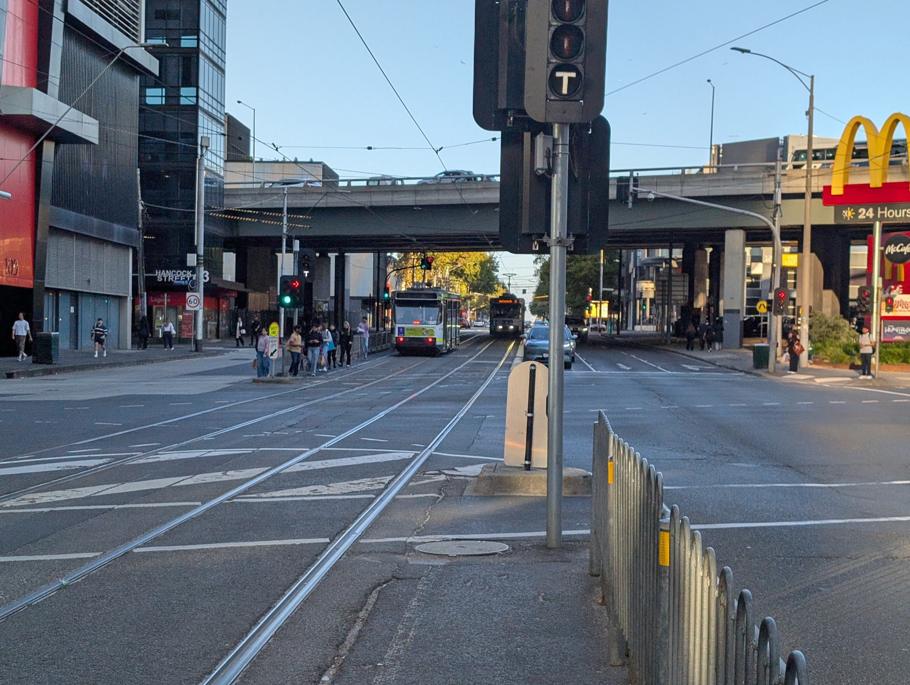 A city street features a tram departing, one approaching, traffic lights, and a mix of pedestrians and modern buildings under a clear sky.