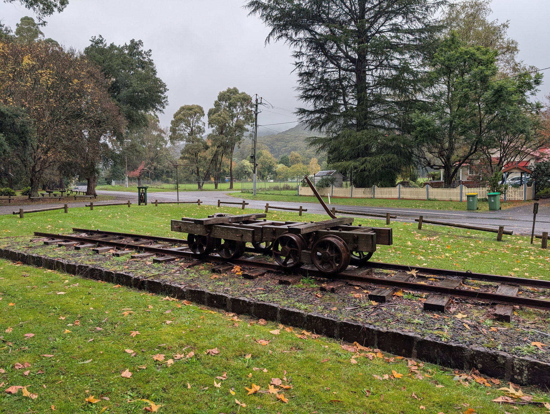 An old, weathered rail cart sits on rusty tracks surrounded by green grass and trees in a park-like setting.