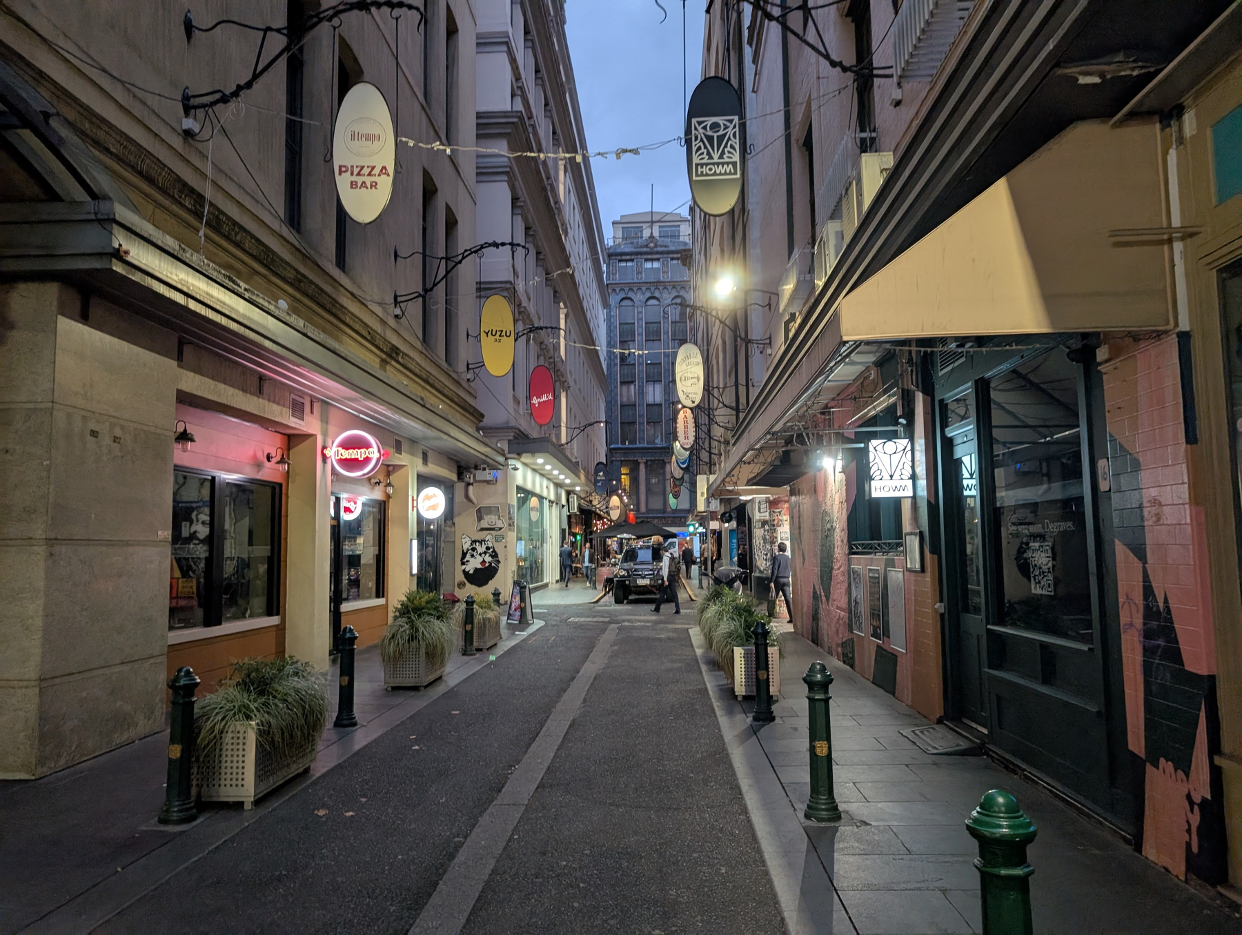 A narrow alley lined with signs and lights leads to a distant building, featuring various shops and restaurants on either side.