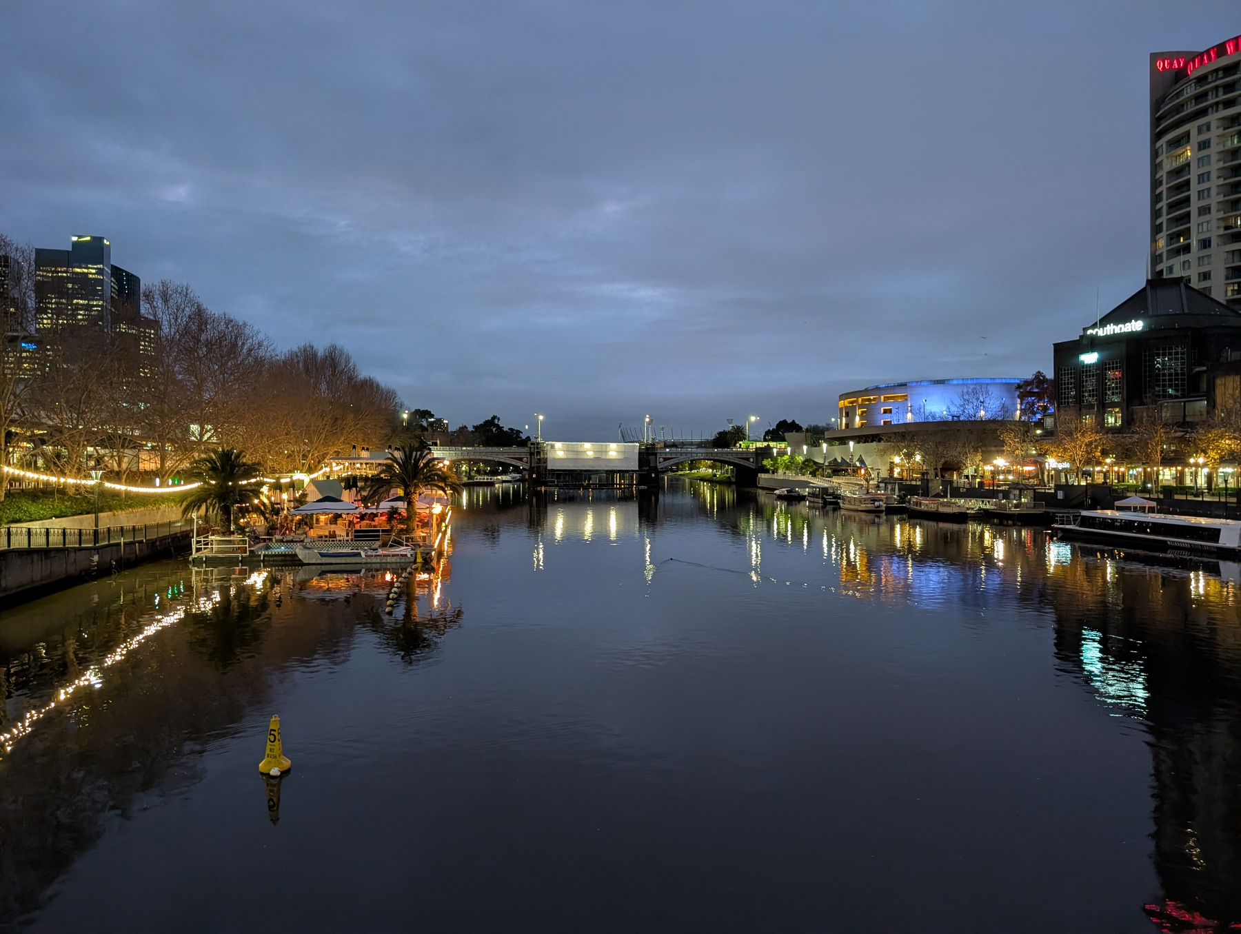A calm river at dusk flows through a cityscape with buildings and illuminated trees reflecting on the water.