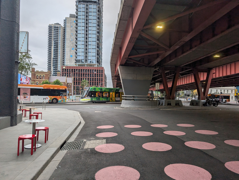 An urban street scene features a tram and bus passing by, tall buildings, pink polka dots on the road, and a bridge overhead.