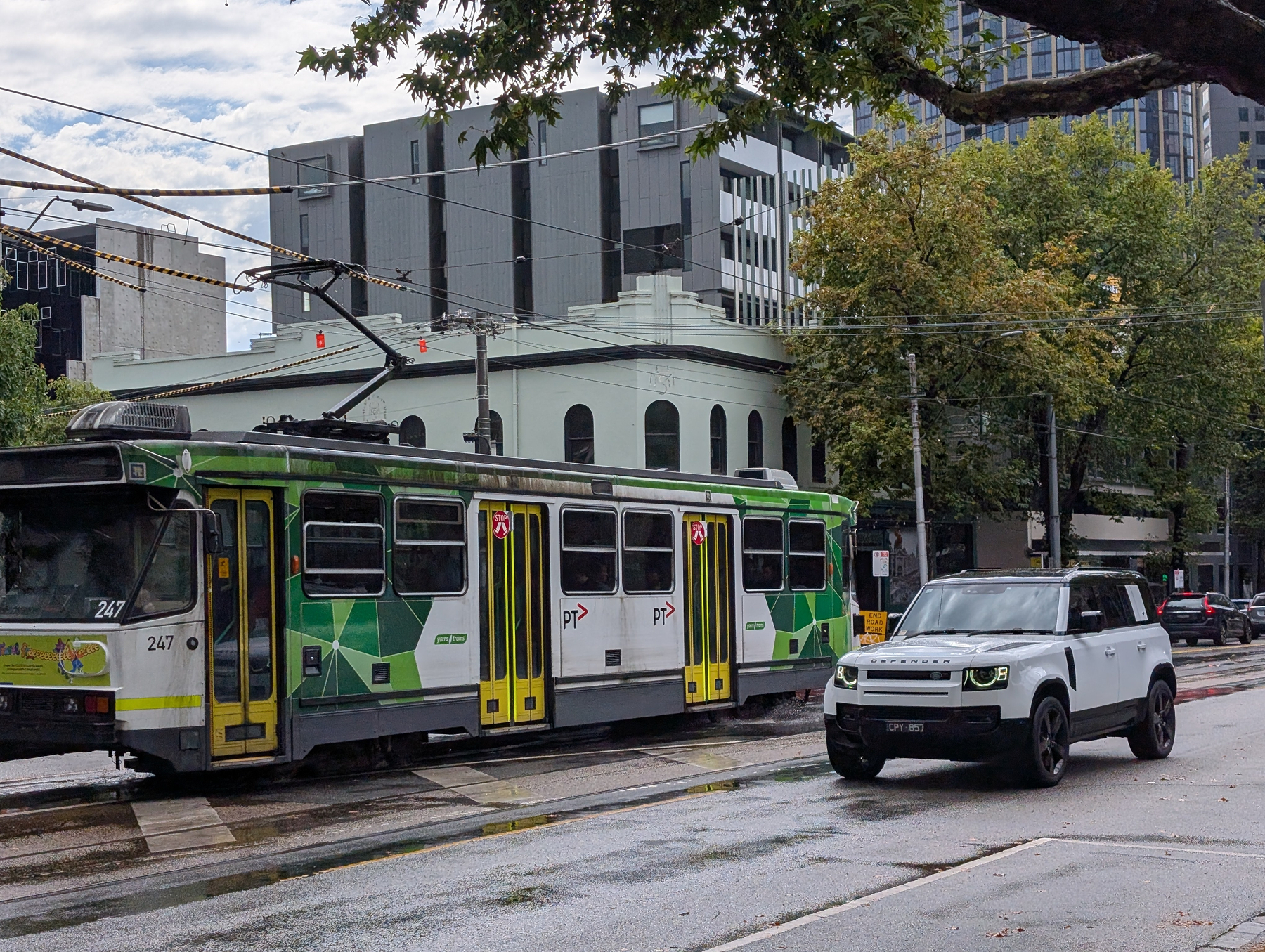 A tram and an SUV are traveling along a city street lined with trees and modern buildings.