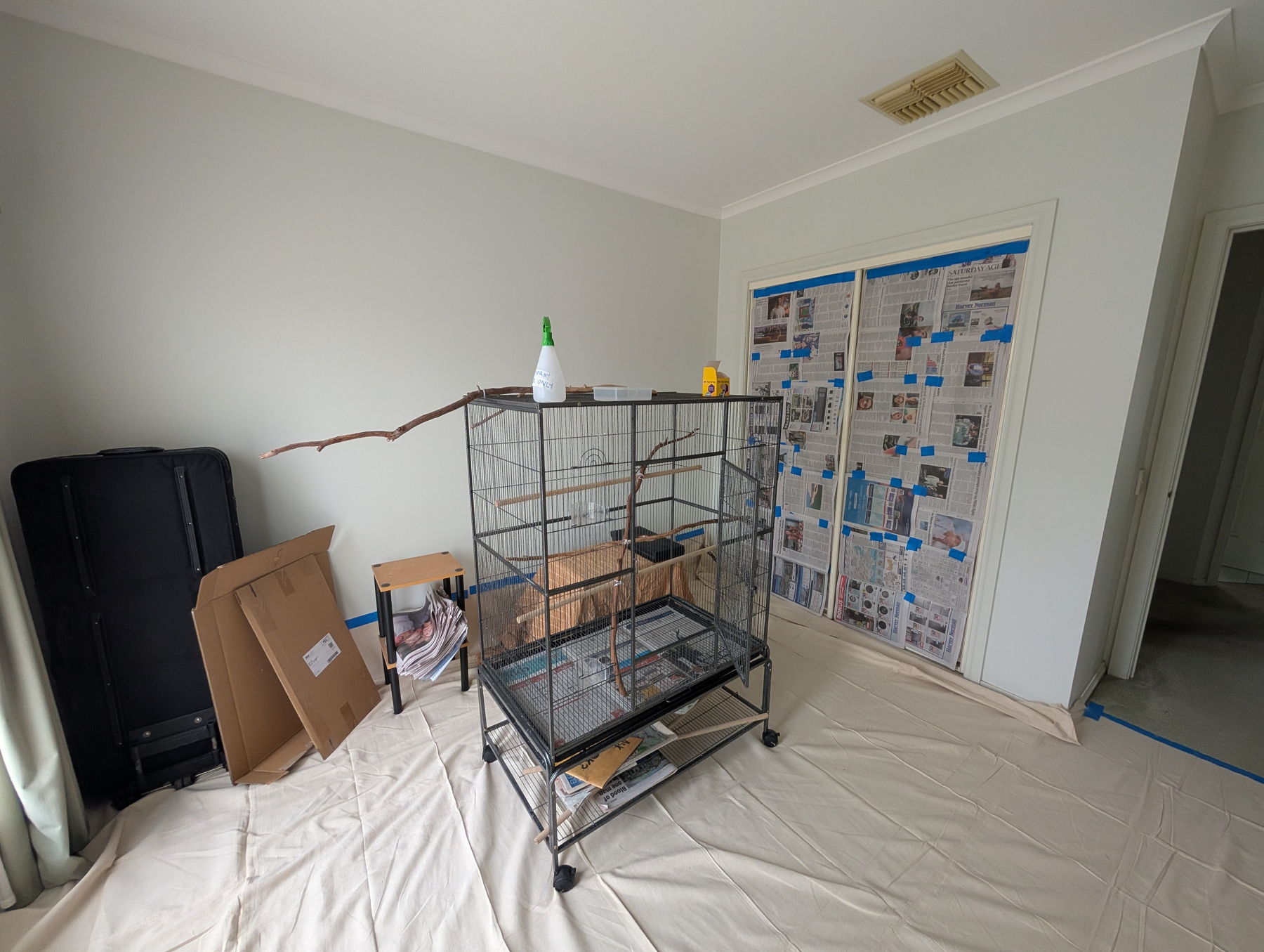 A room with a large birdcage on wheels surrounded by folded cardboard boxes, a small stool, and protective coverings on the floor and doorway, suggesting preparation for painting or renovation.