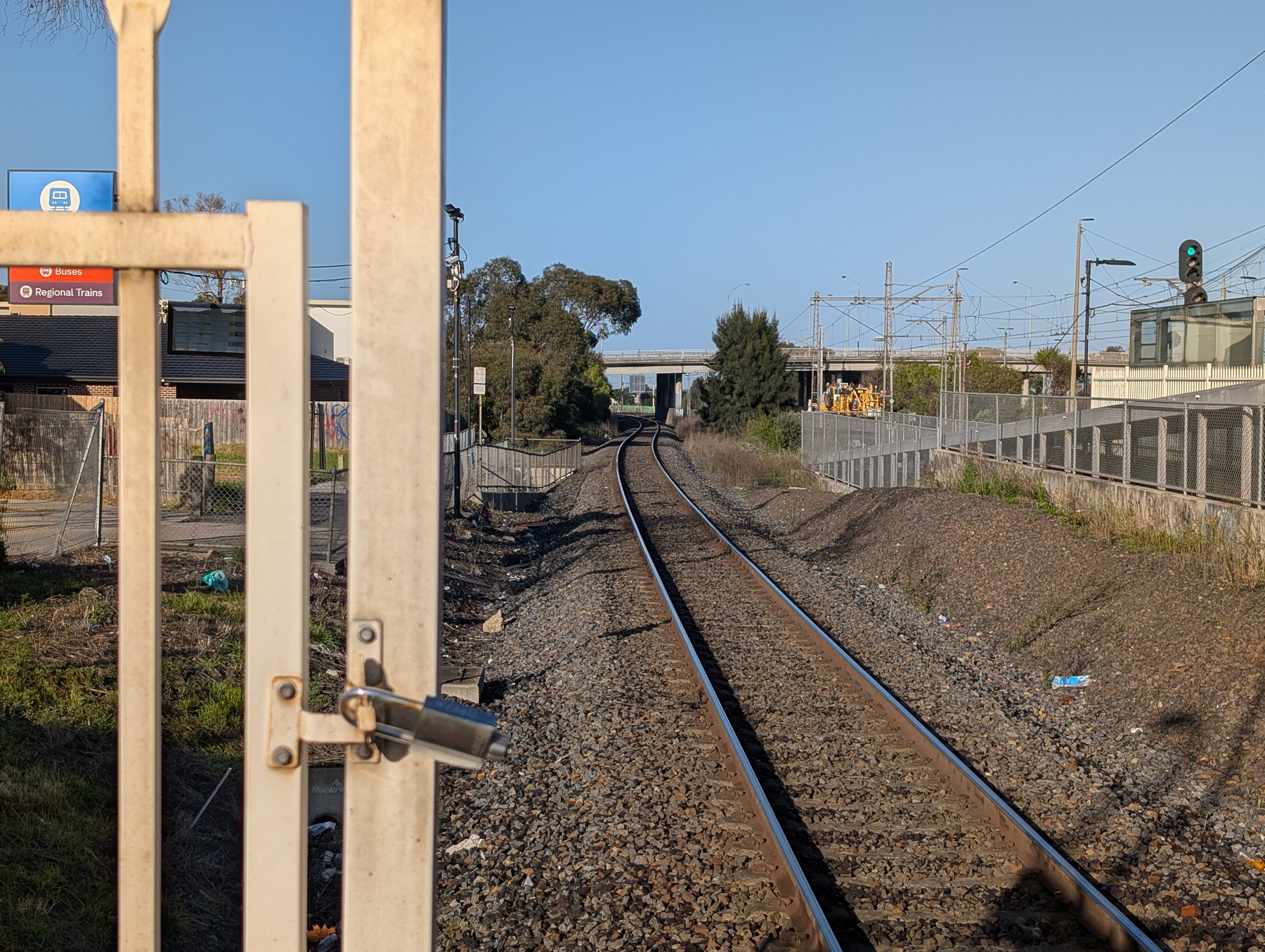 Railway tracks stretch into the distance beside a fenced area and a small train station under a clear blue sky.