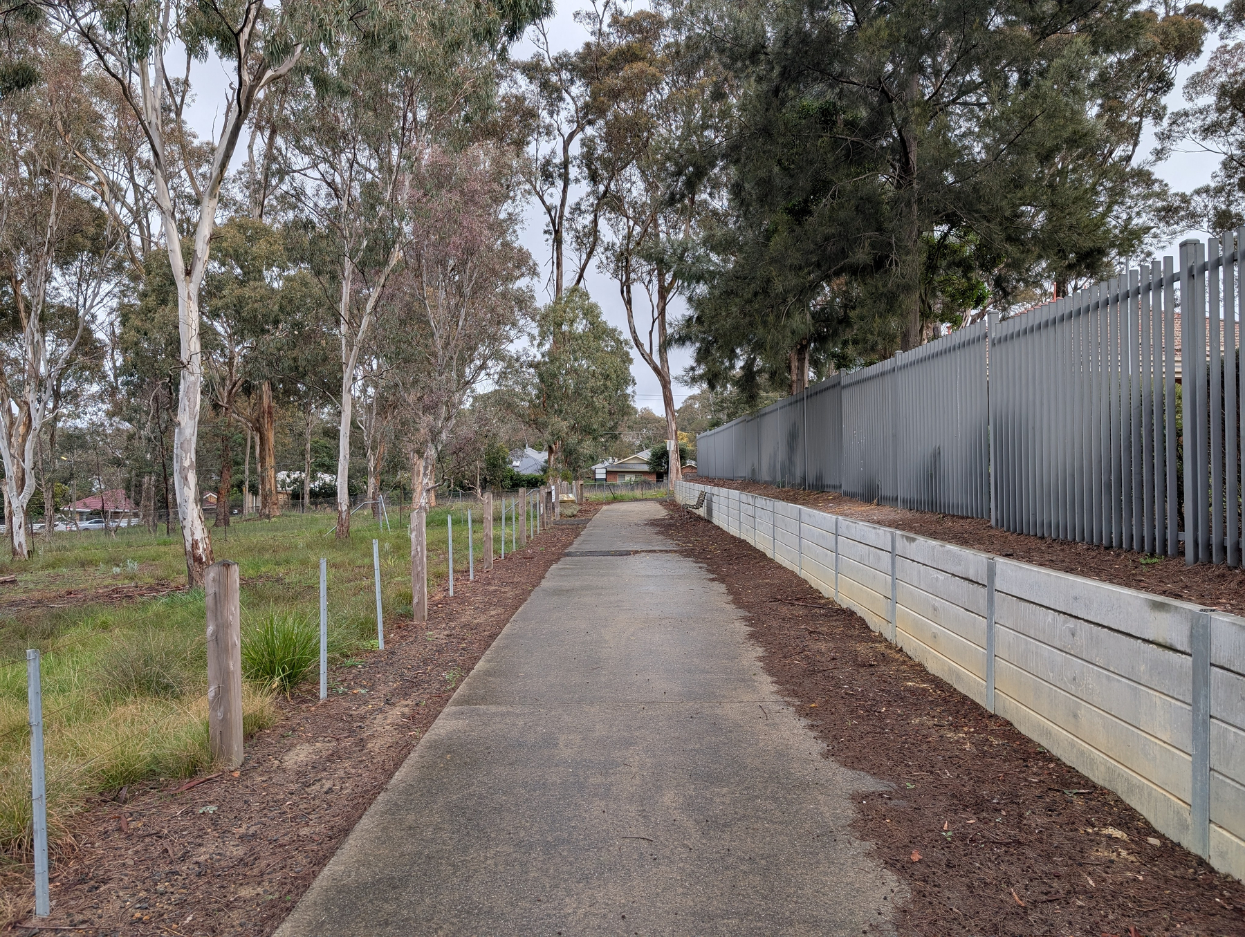 A paved pathway runs between a grassy area with trees on the left and a fenced area with a retaining wall on the right.
