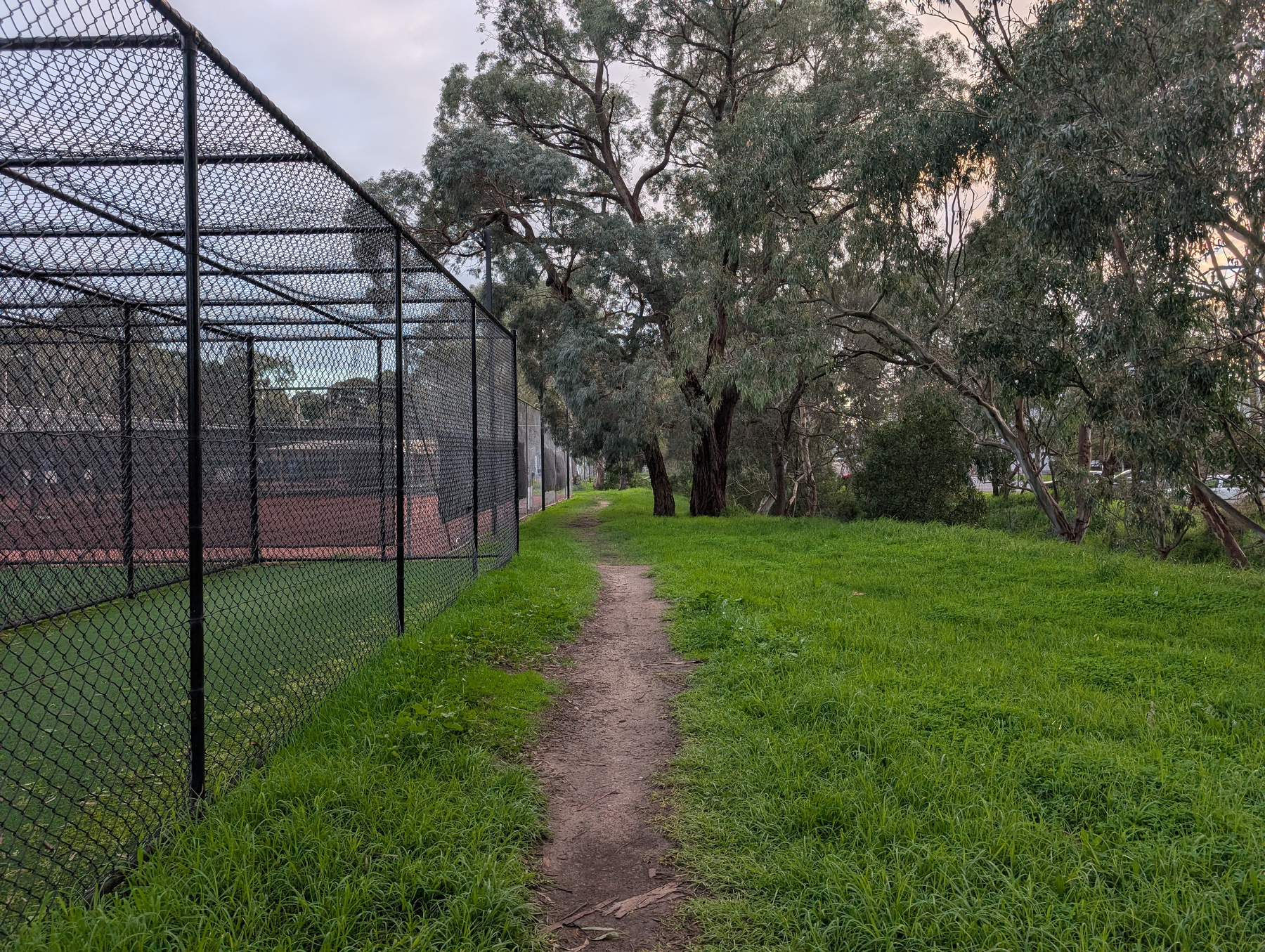 Auto-generated description: A narrow dirt path runs alongside a fenced tennis court with green grass and tall trees on the other side.
