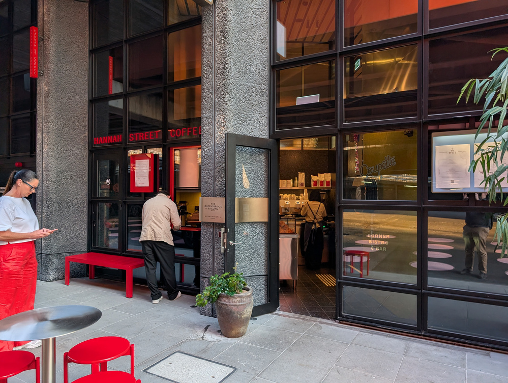 A woman in a red skirt looks at her phone outside a coffee shop with a glass facade, while a man stands at the counter inside.