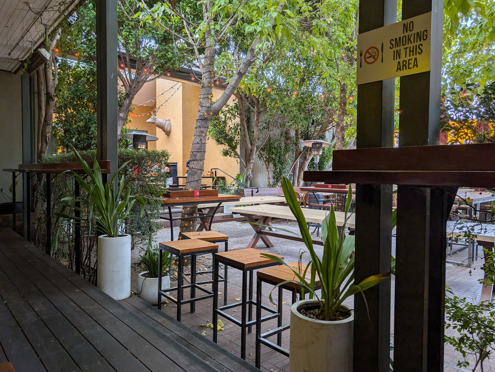 A cozy outdoor seating area features wooden tables and stools, surrounded by lush greenery and a No Smoking sign.