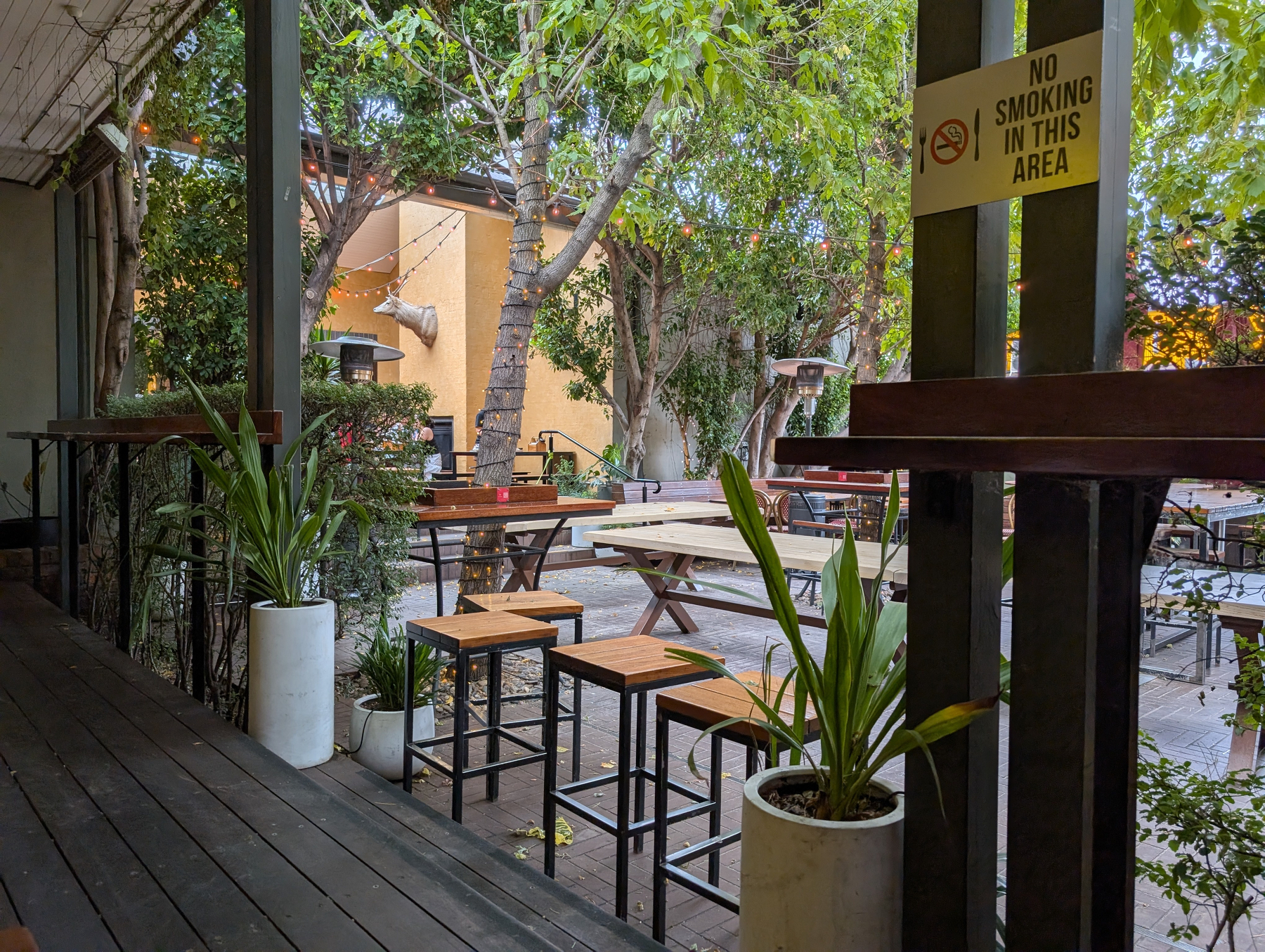 Auto-generated description: A cozy outdoor seating area features wooden tables and stools, surrounded by lush greenery and a No Smoking sign.