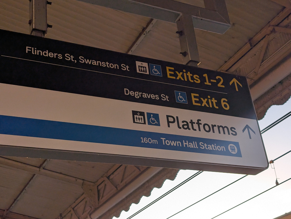 A directional sign at a train station points towards exits 1-2, exit 6, and platforms, with specific references to Flinders St, Swanston St, Degraves St, and Town Hall Station.