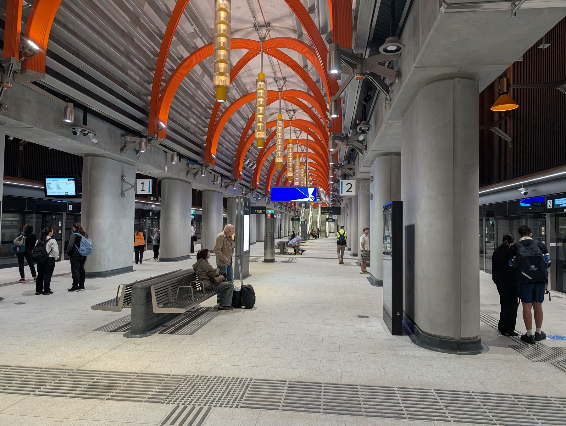 Auto-generated description: A modern underground station features orange arching beams, signage, and people waiting or walking within the spacious platform area.