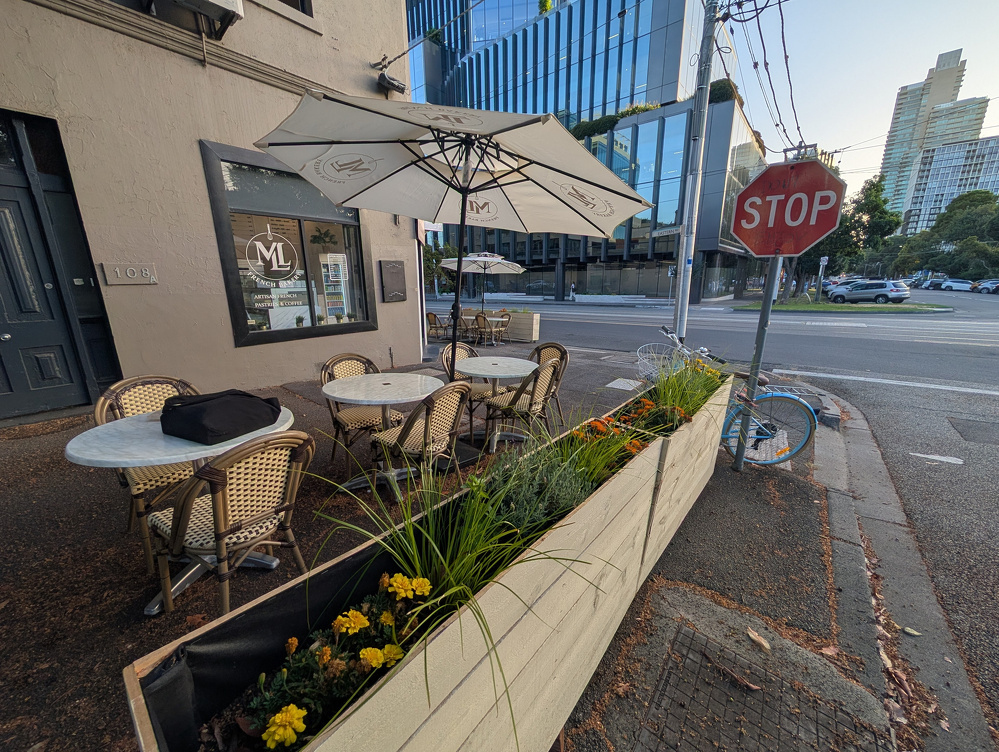 A sidewalk café features tables with chairs and umbrellas next to a planter with yellow flowers, situated near a modern building and a stop sign.