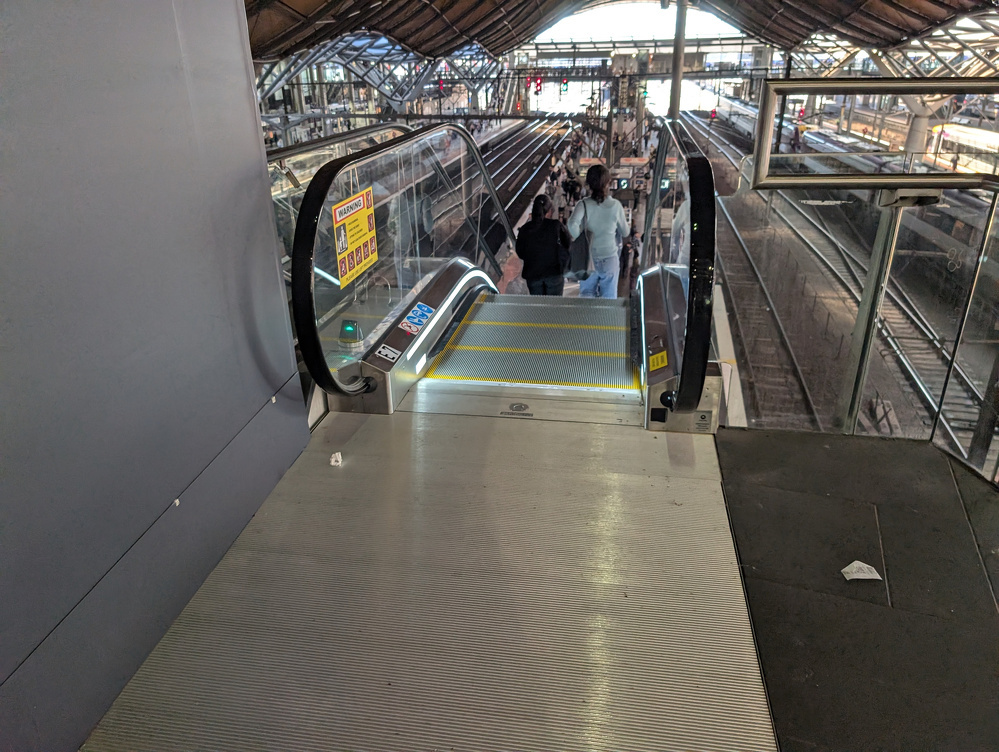 An escalator leads down to a train station platform, with people standing near the tracks under a large, industrial-style roof.