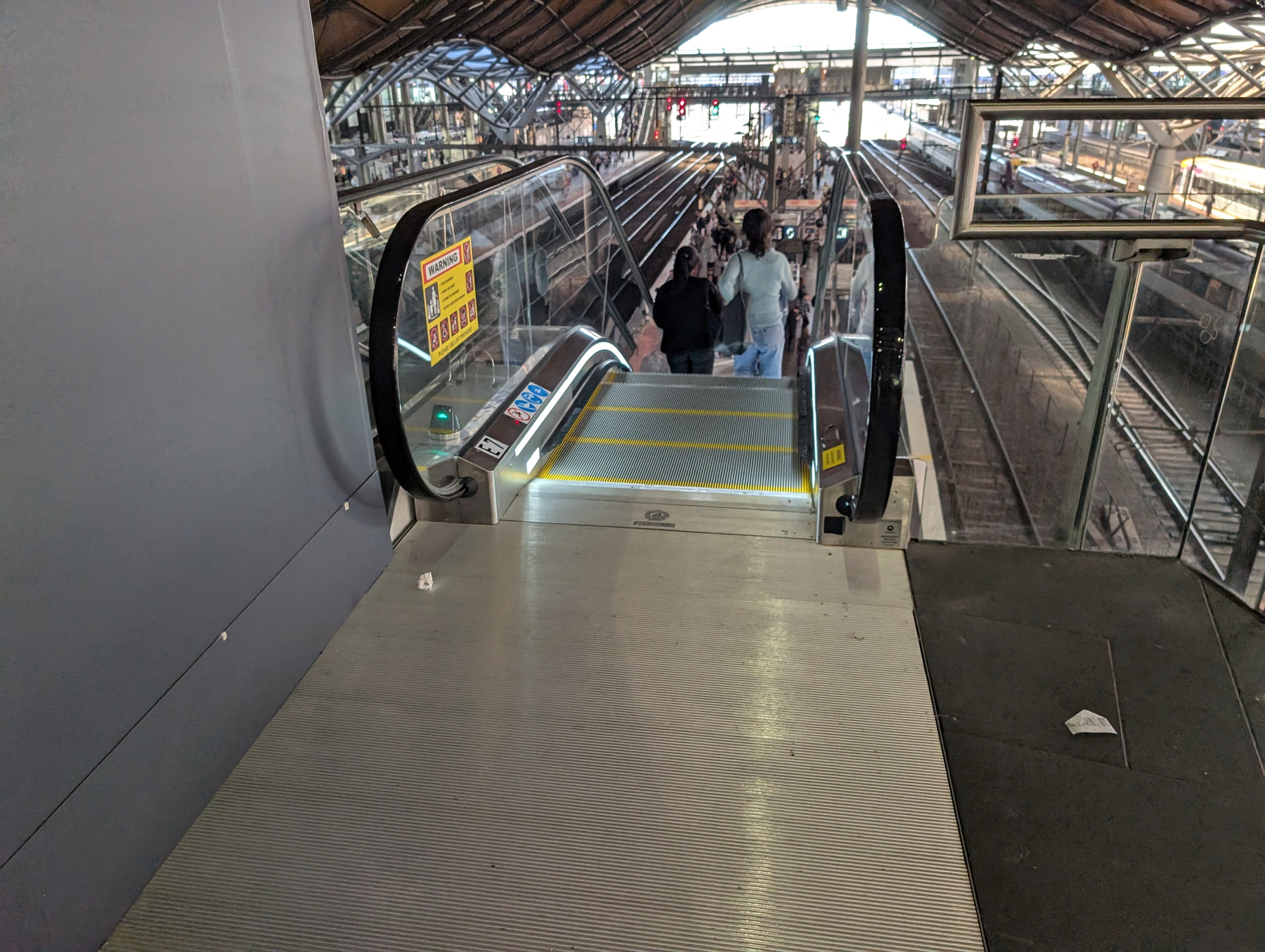 Auto-generated description: An escalator leads down to a train station platform, with people standing near the tracks under a large, industrial-style roof.