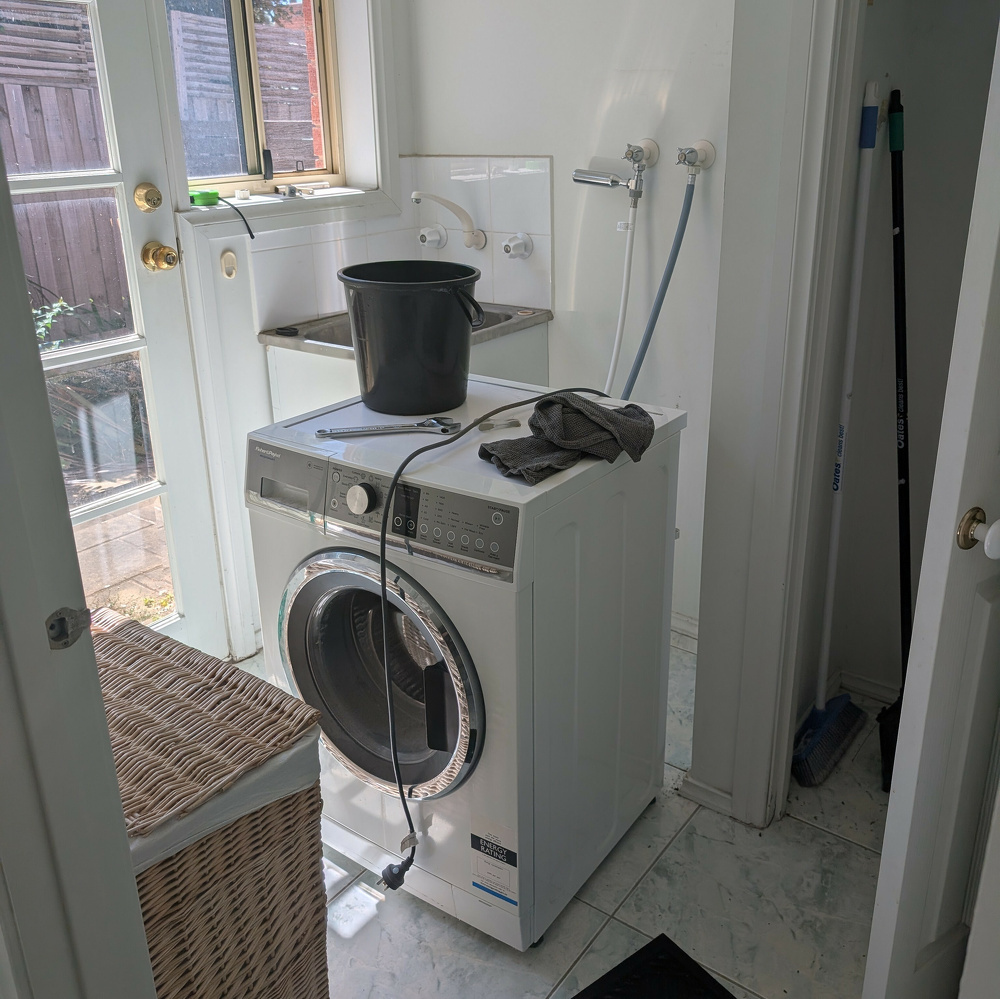A laundry room contains a washing machine with a black bucket on top, various cleaning tools, and access to a sunny outdoor area through a door and window.