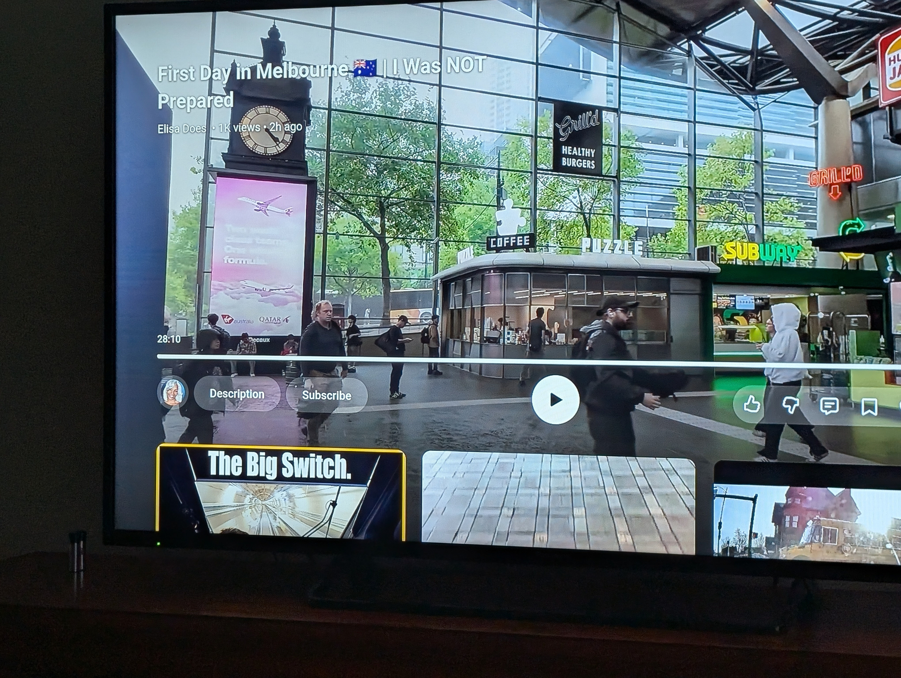 Auto-generated description: A television screen displays a YouTube video titled First Day In Melbourne π¦πΊ I Was NOT Prepared, featuring a city scene with people and shops.