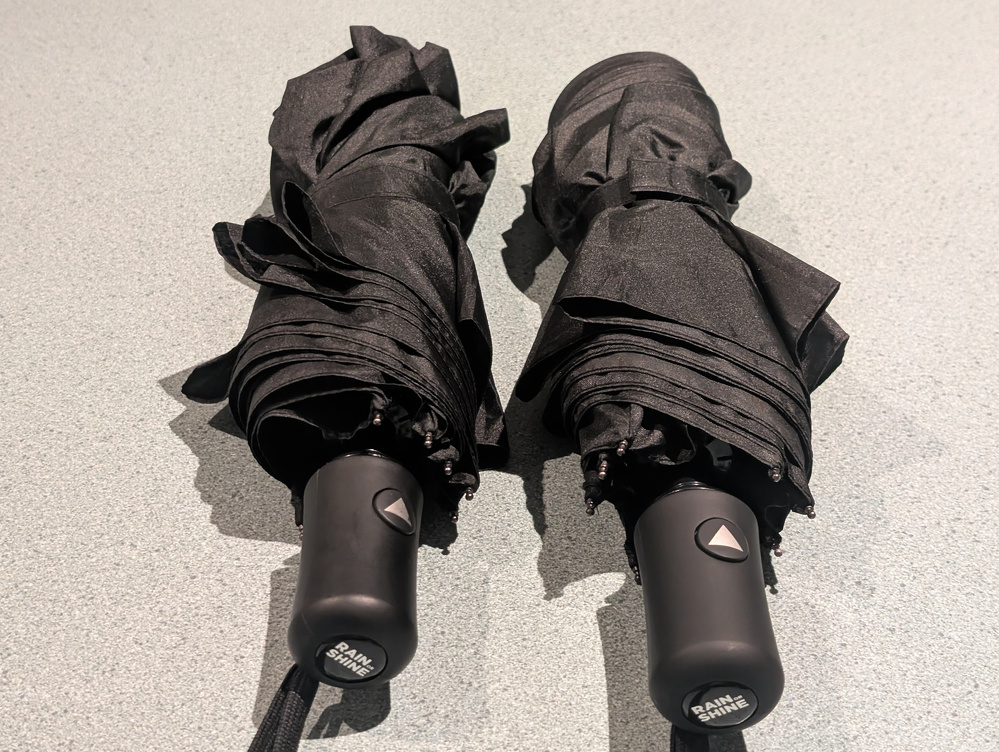 Two black, compact umbrellas with automatic open buttons are placed side by side on a light-colored surface.