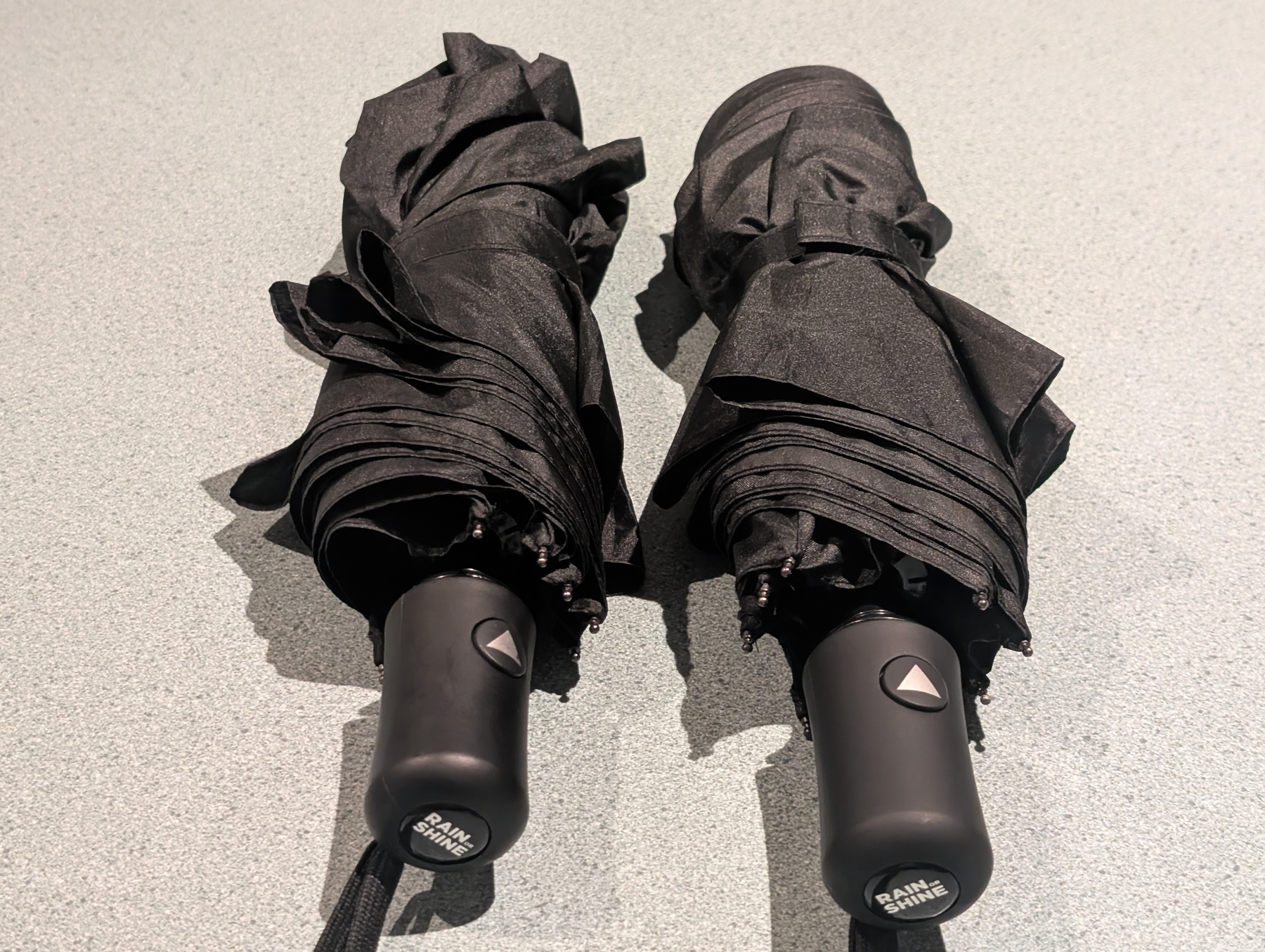 Two black, compact umbrellas with automatic open buttons are placed side by side on a light-colored surface.