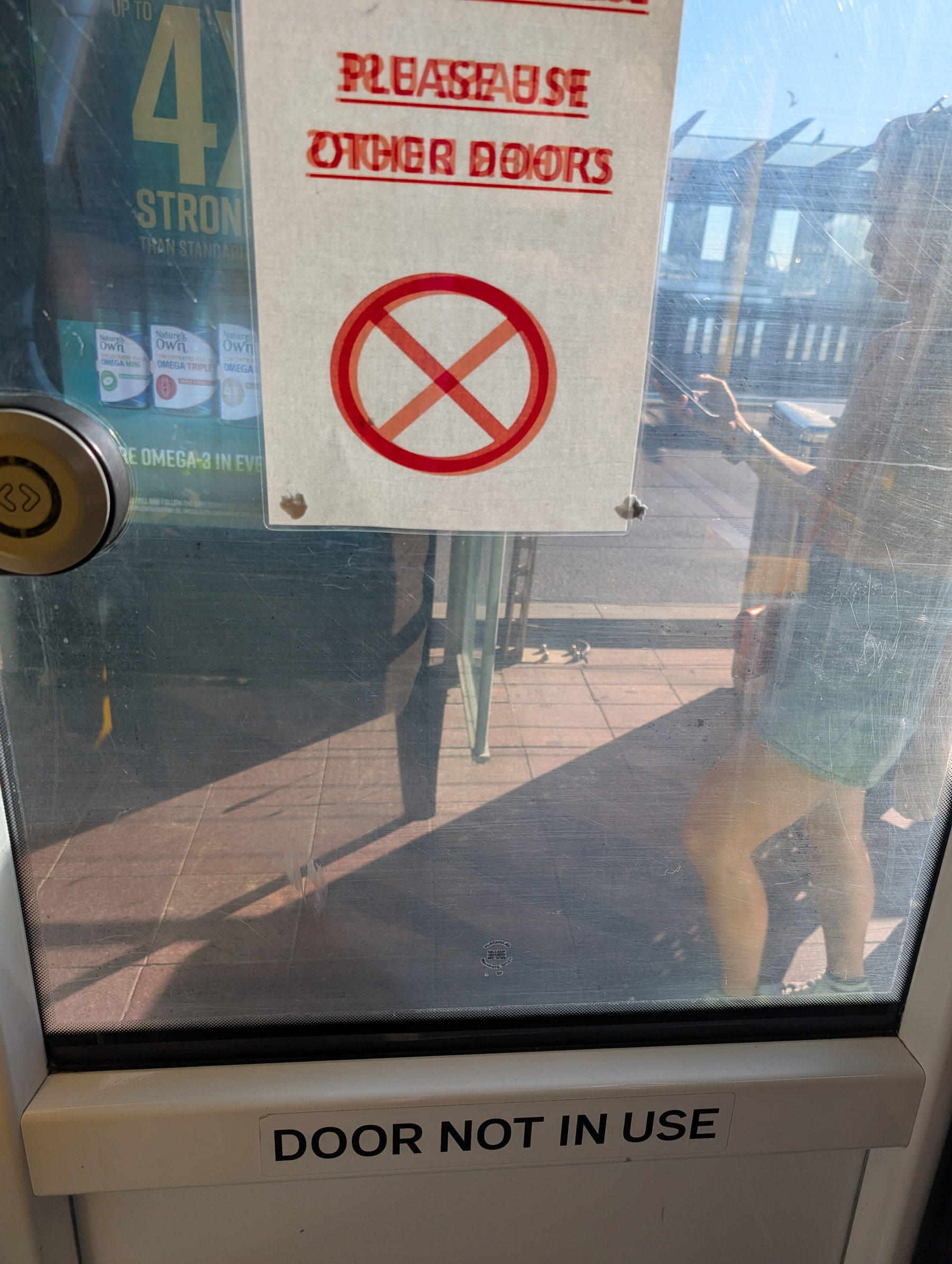 A door with a sign saying PLEASE USE OTHER DOOR and DOOR NOT IN USE is situated in front of an outside area with people.