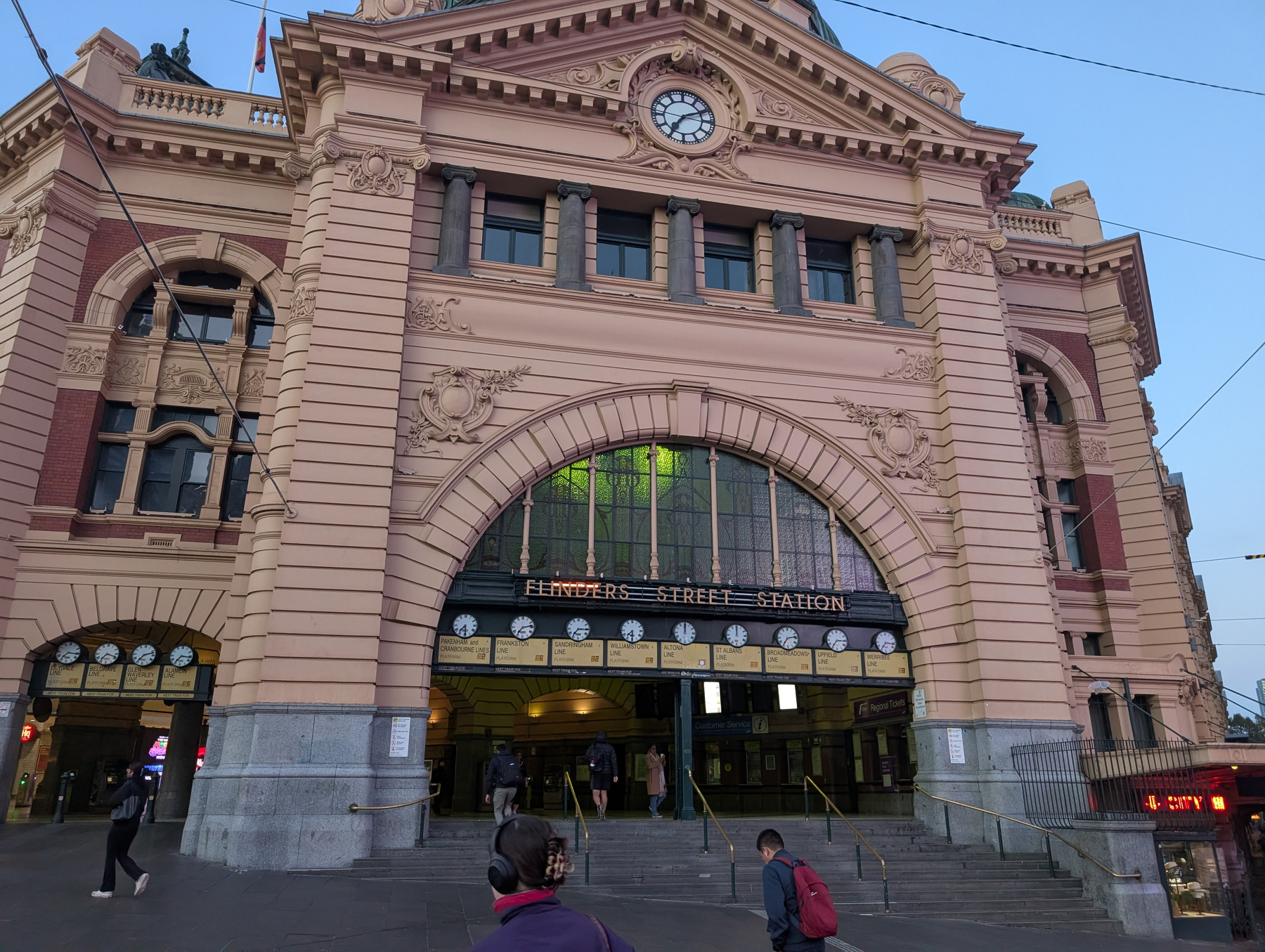 Auto-generated description: People are entering and exiting Flinders Street Station, an iconic historic building in Melbourne, Australia, with its distinctive arched facade and clock.