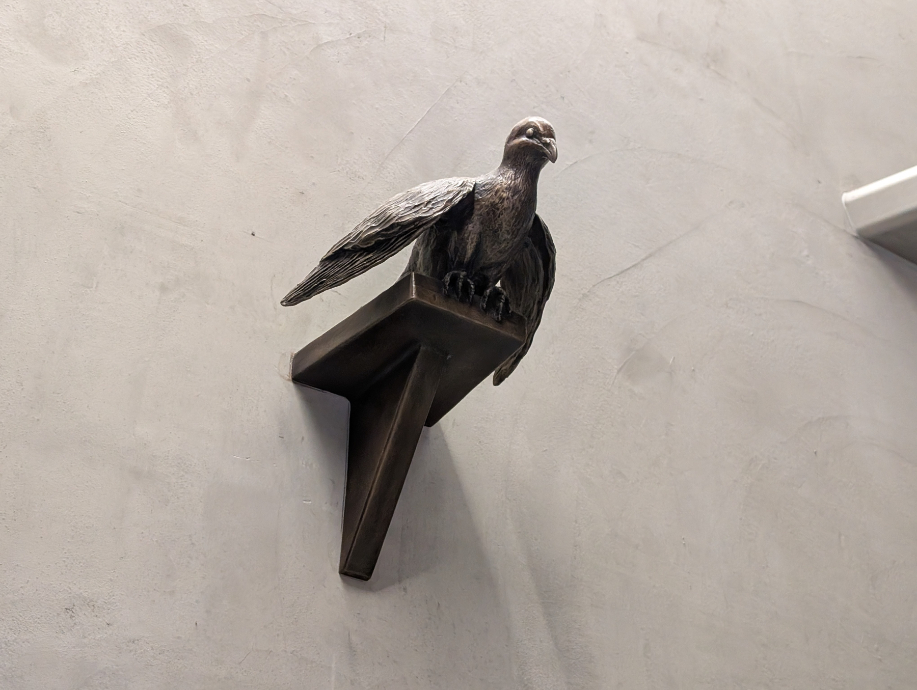 Auto-generated description: A stone sculpture of a bird is perched on a triangular shelf protruding from a textured wall.