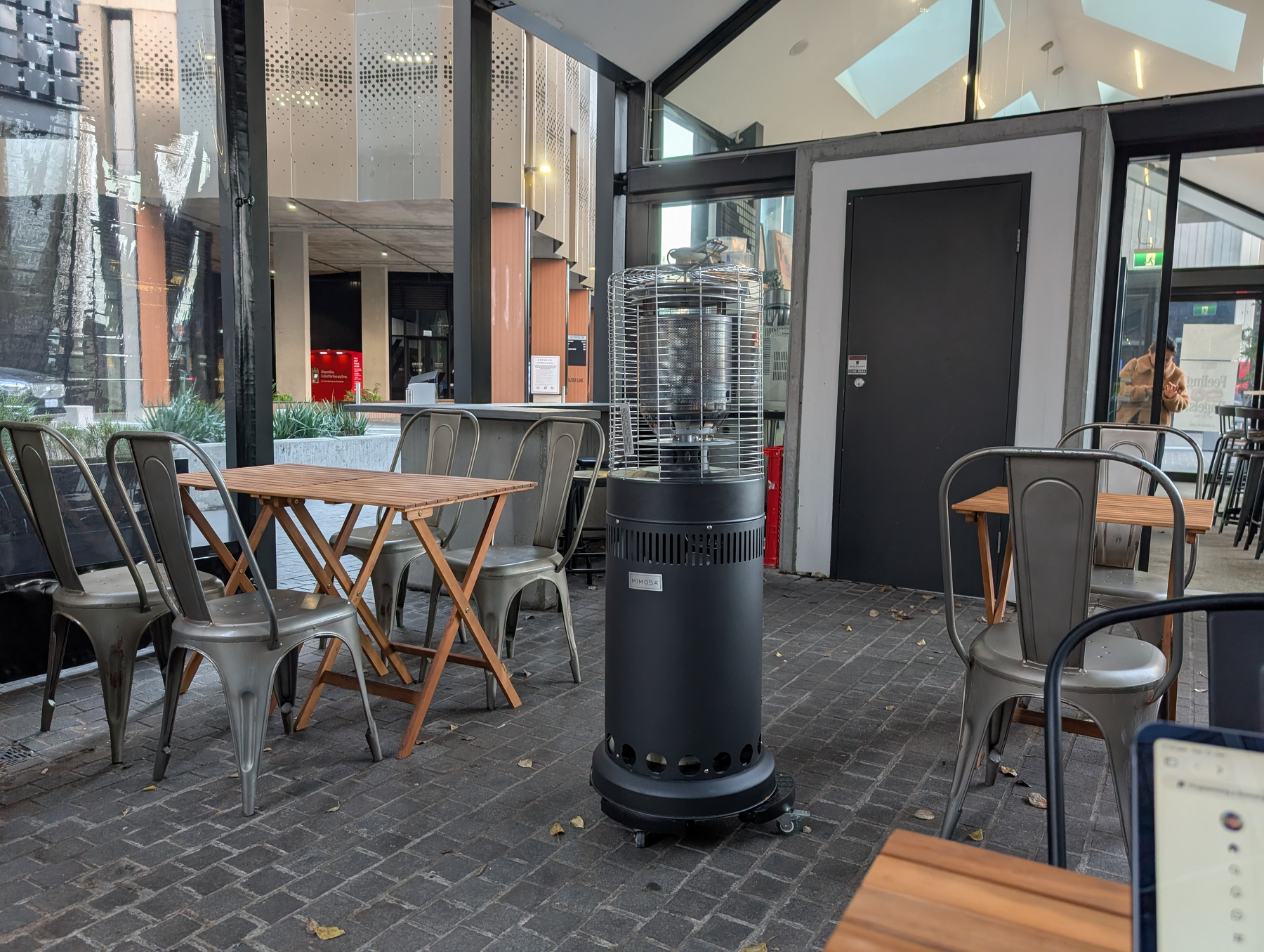 Auto-generated description: A cafe seating area features empty metal chairs and wooden tables, with a portable heater in the center.
