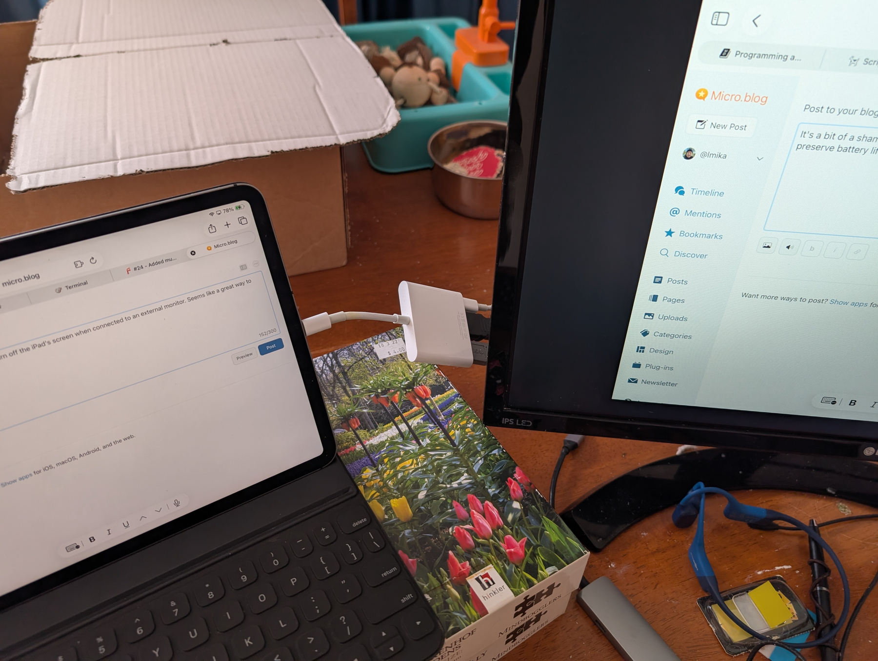 Auto-generated description: Two screens are displaying text on a wooden desk cluttered with various items, including a box, a tangled cord, and vibrant pencils.