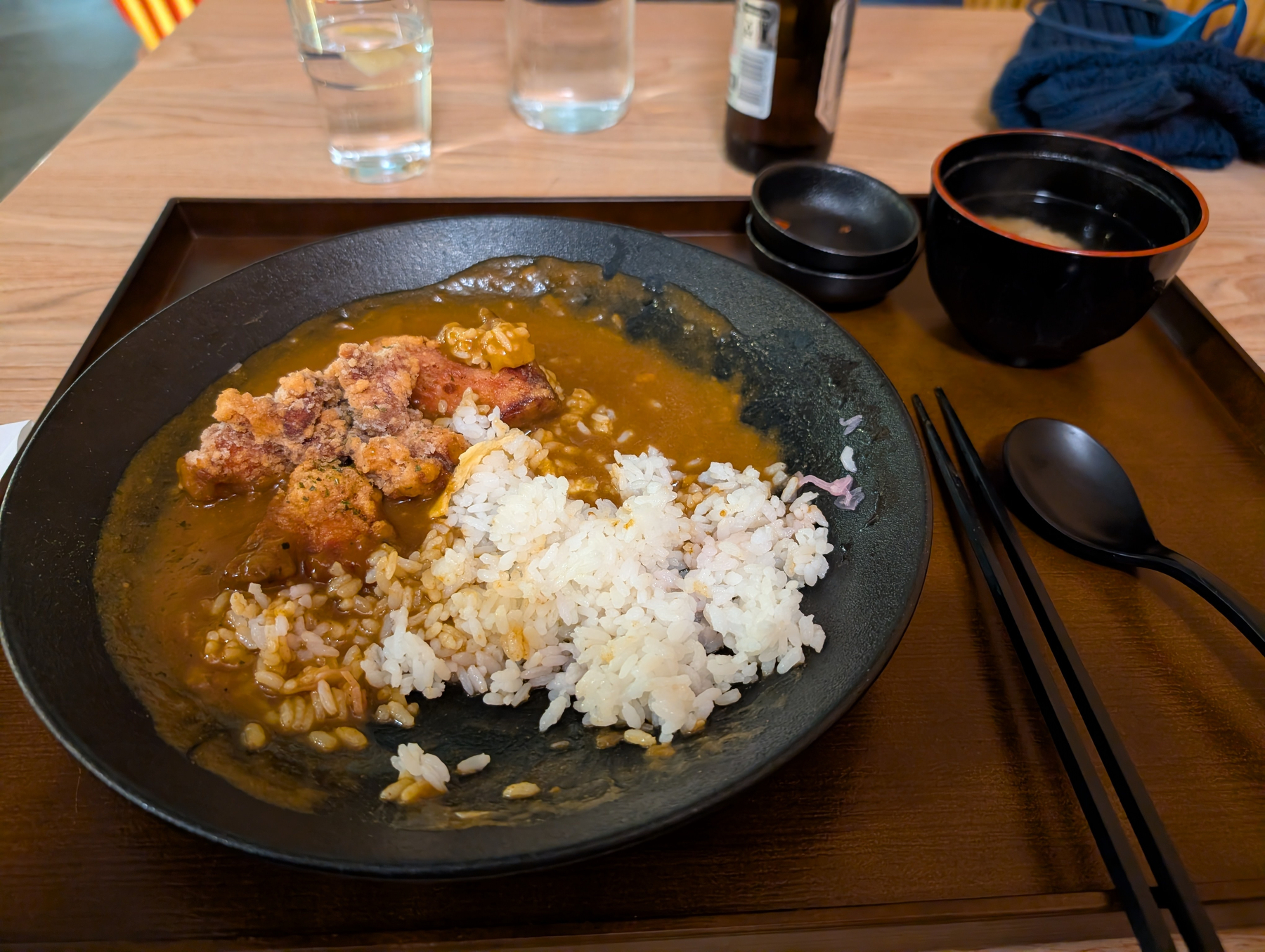 Auto-generated description: A plate of Japanese curry with rice and fried chicken is served on a tray along with a bowl of soup, chopsticks, a spoon, and two glasses of water on a wooden table.